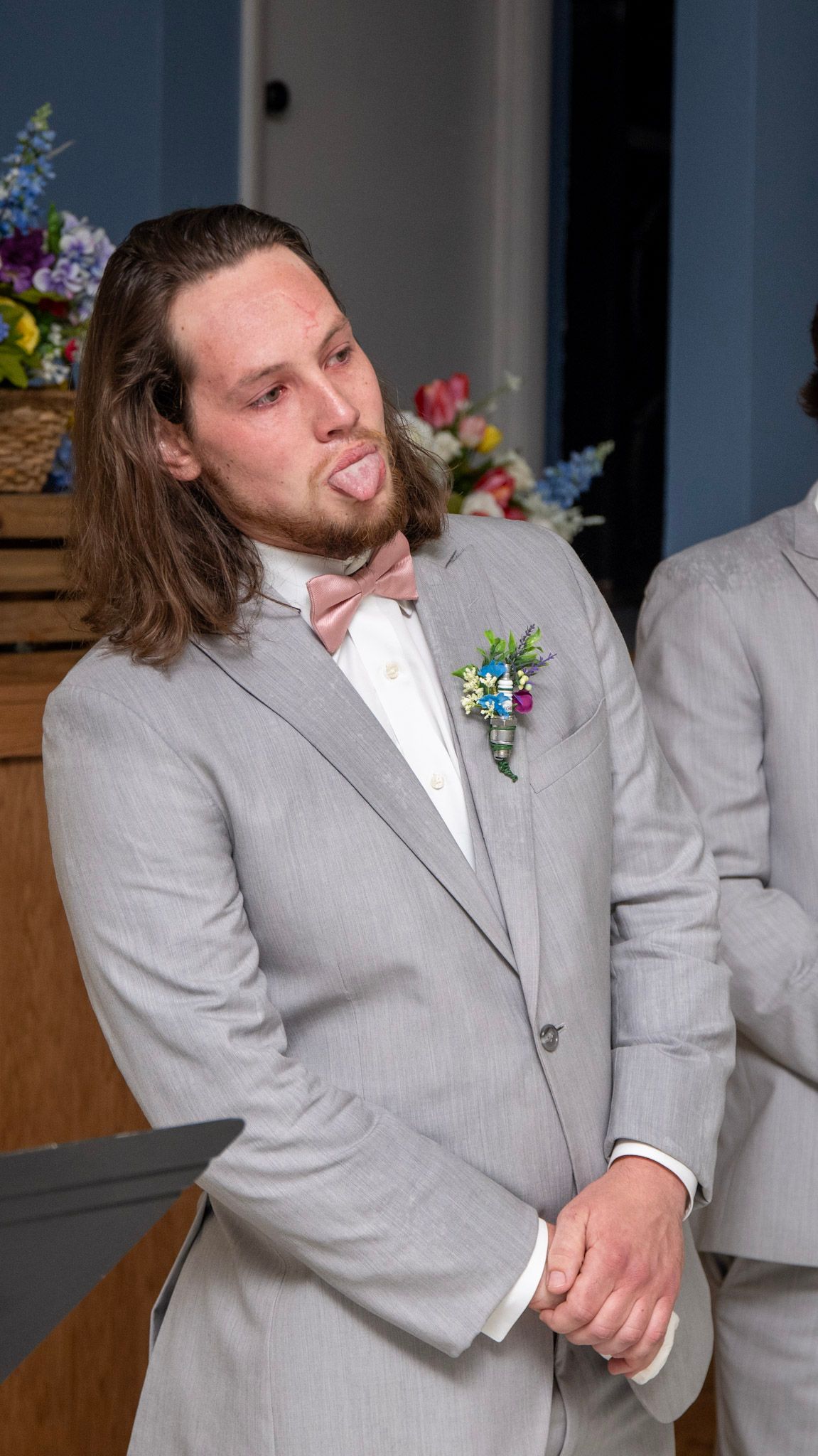 Groomsman in light gray suit and white boutonniere shows expression during wedding ceremony.