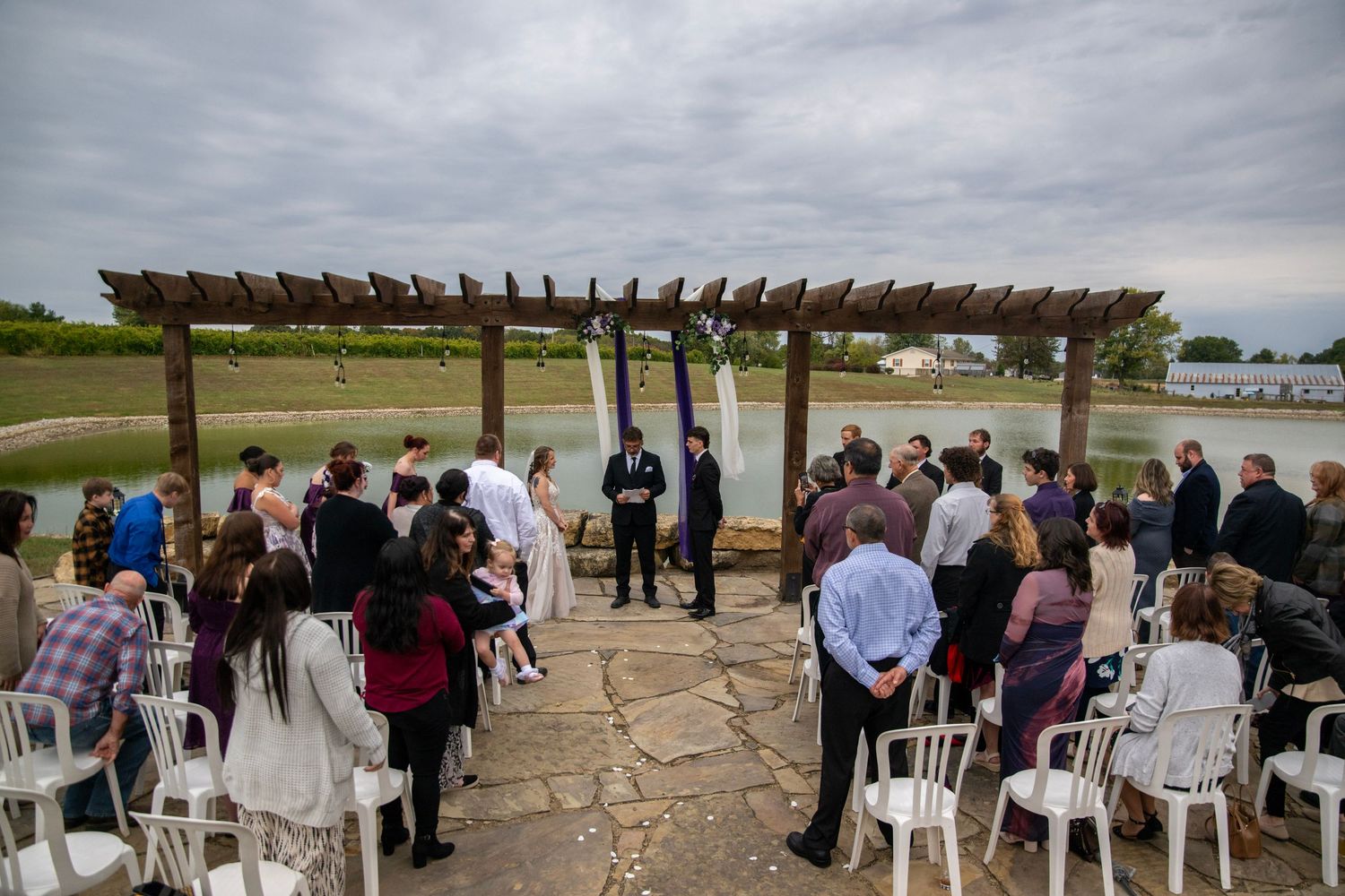 Wide view of wedding ceremony setup with white chairs and lakeside pergola.
