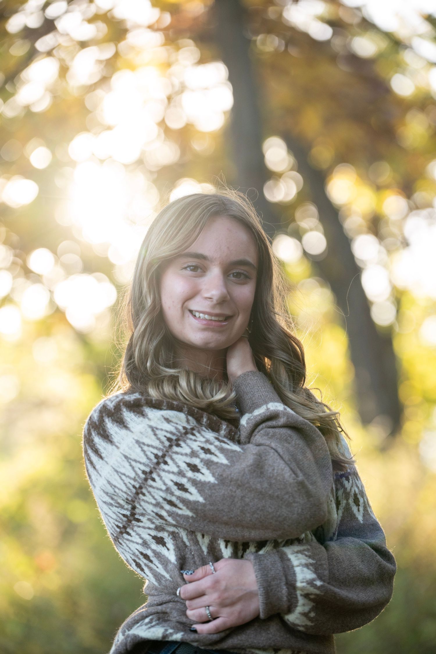 A person wearing a gray patterned sweater poses in a sunlit forest setting with golden bokeh in the background.