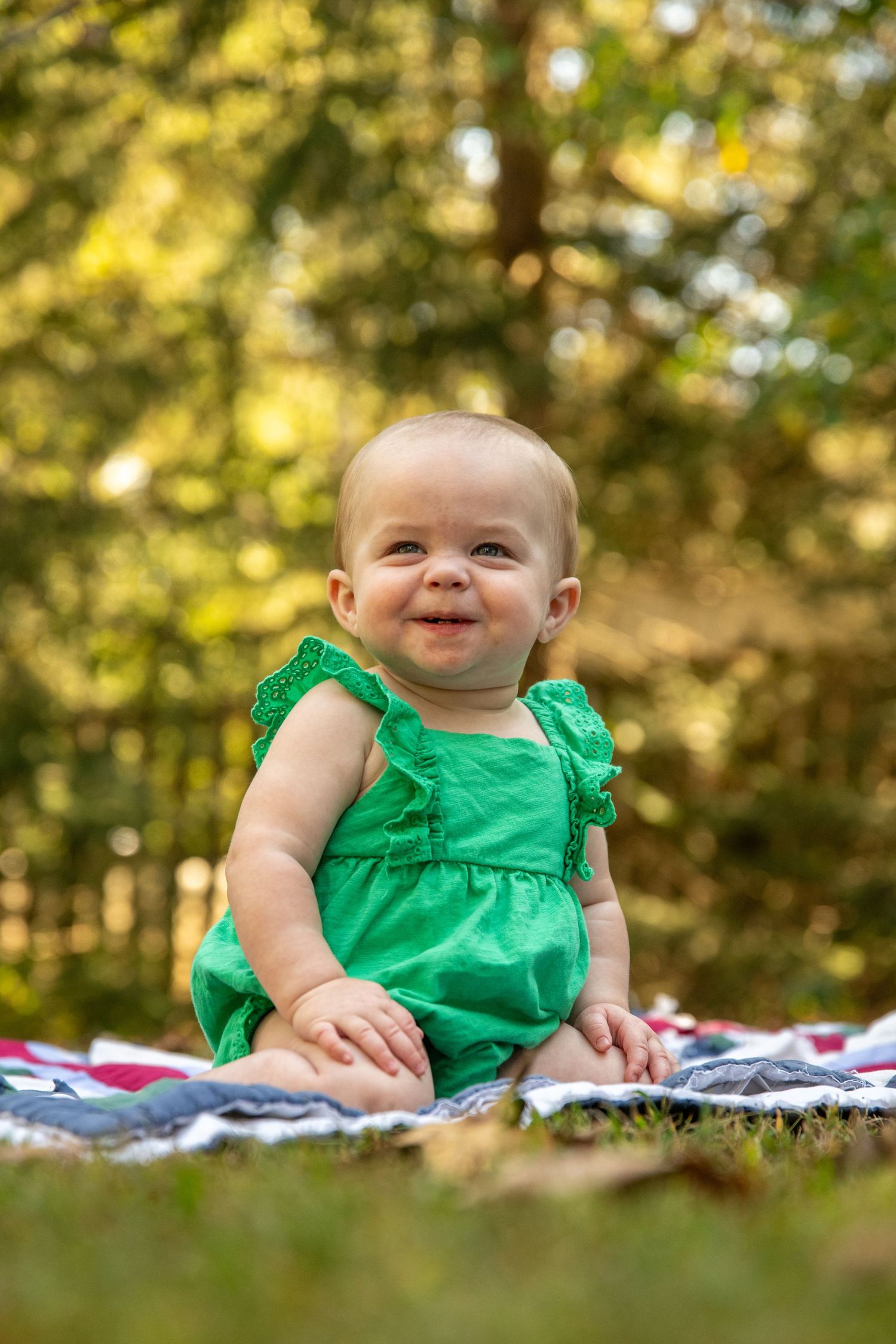 Baby wearing ruffled green dress smiling while sitting on patterned quilt outdoors.