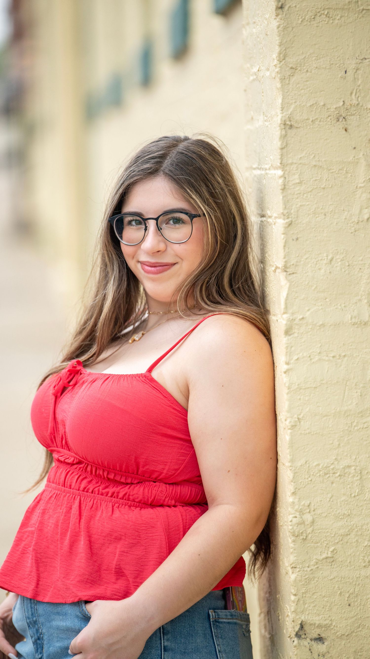 A person in glasses leans against a yellow wall wearing a red top and jeans.