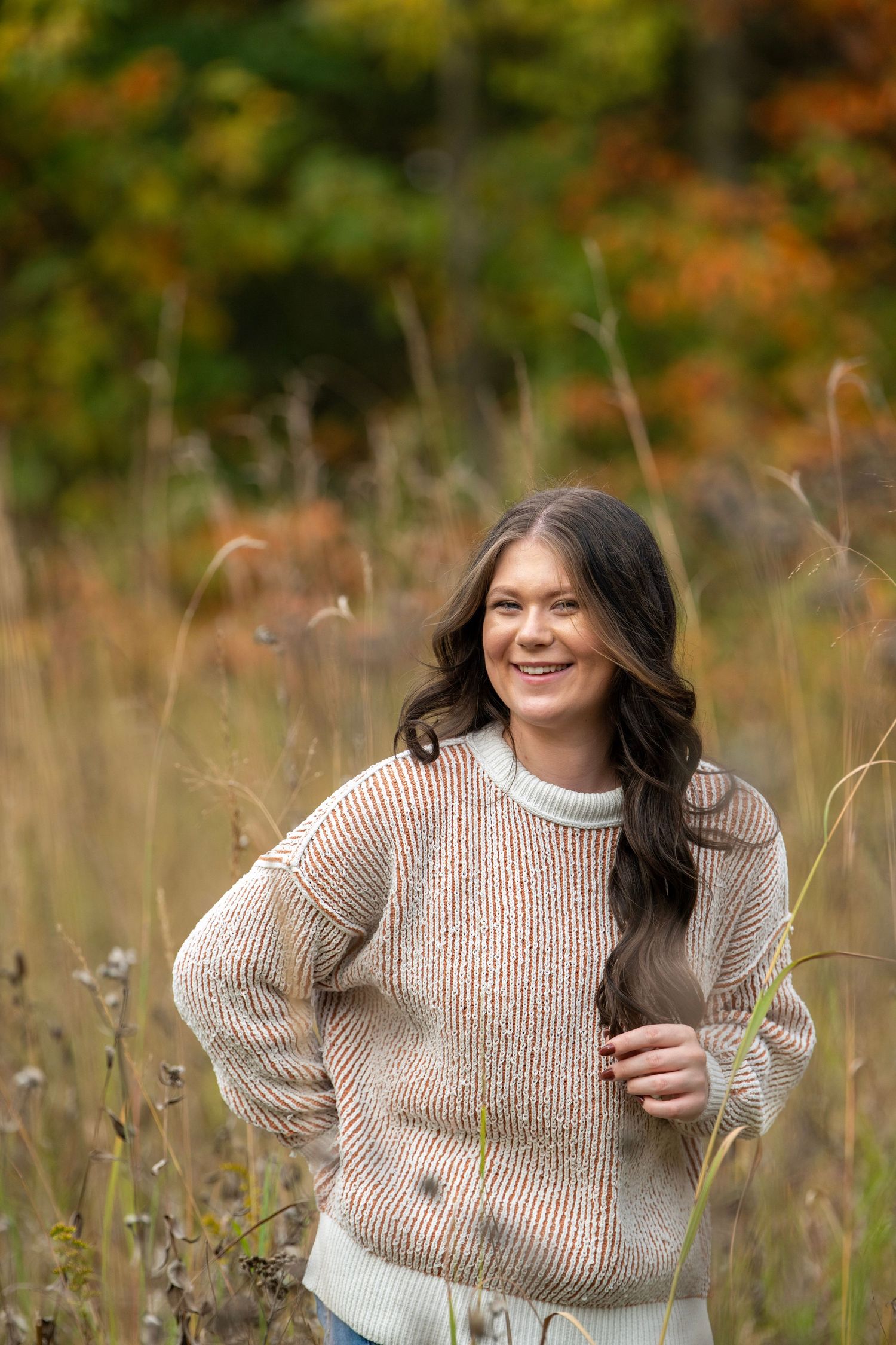 Person in beige sweater smiling while standing in tall grass with autumn colors behind.