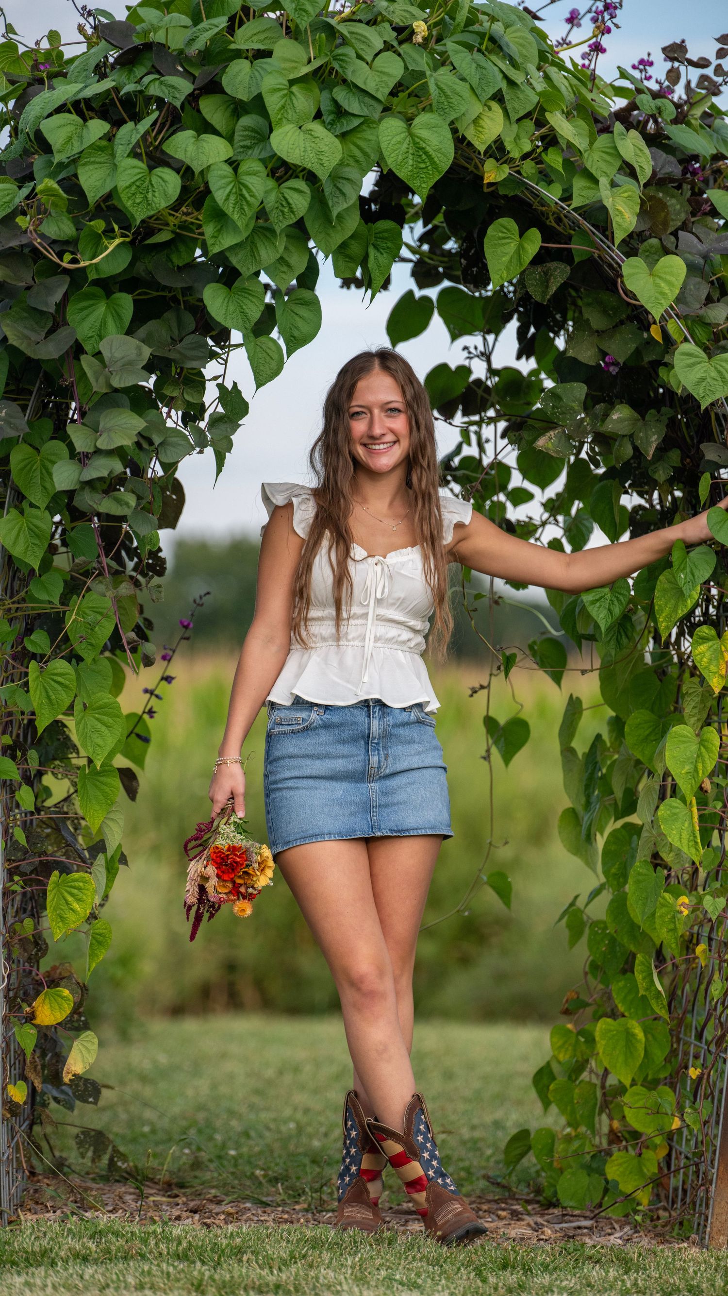 A person poses under a leafy garden arch wearing a white top and denim skirt.