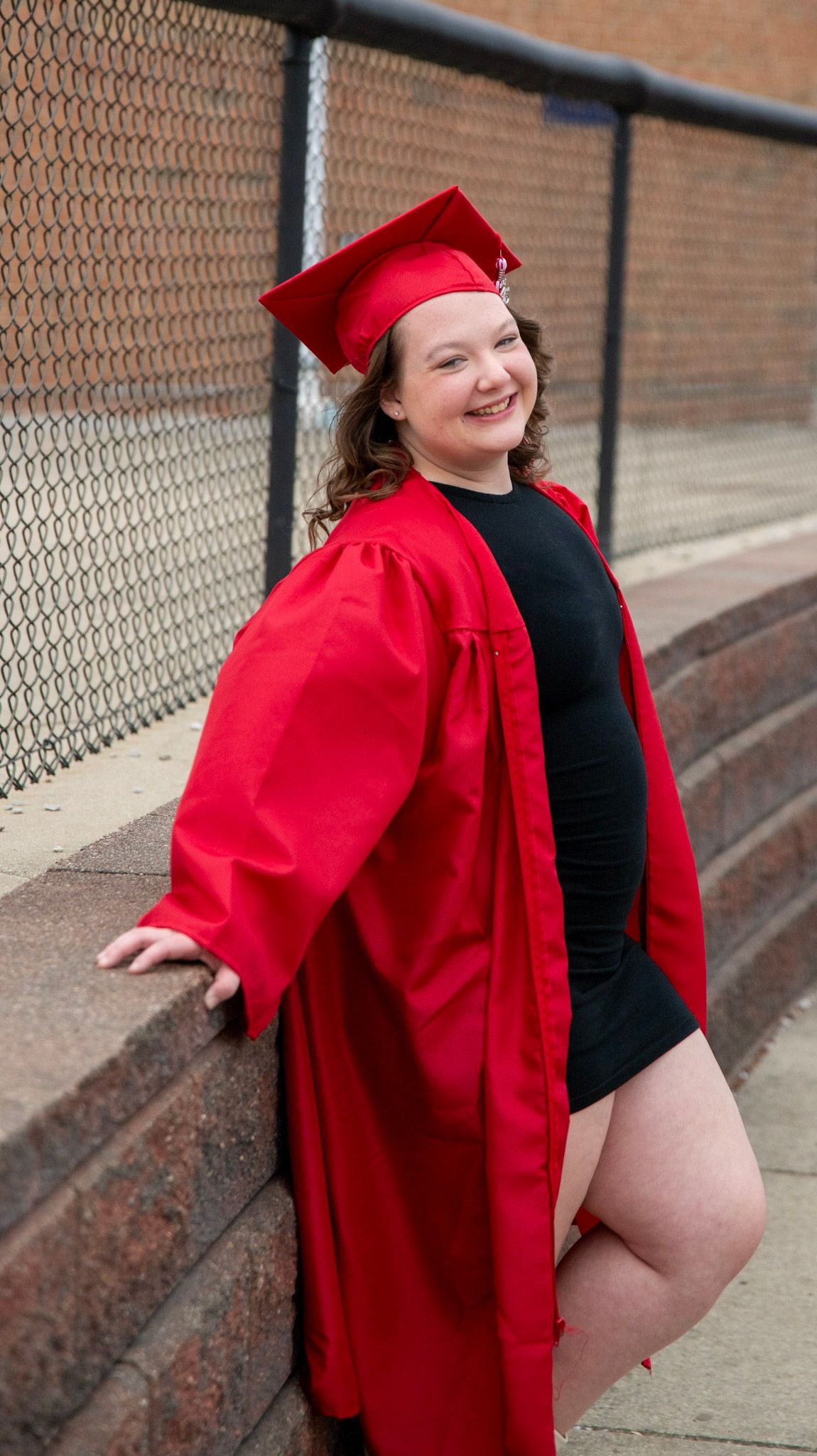 Graduate in bright red graduation attire poses near chain link fence and brick wall.
