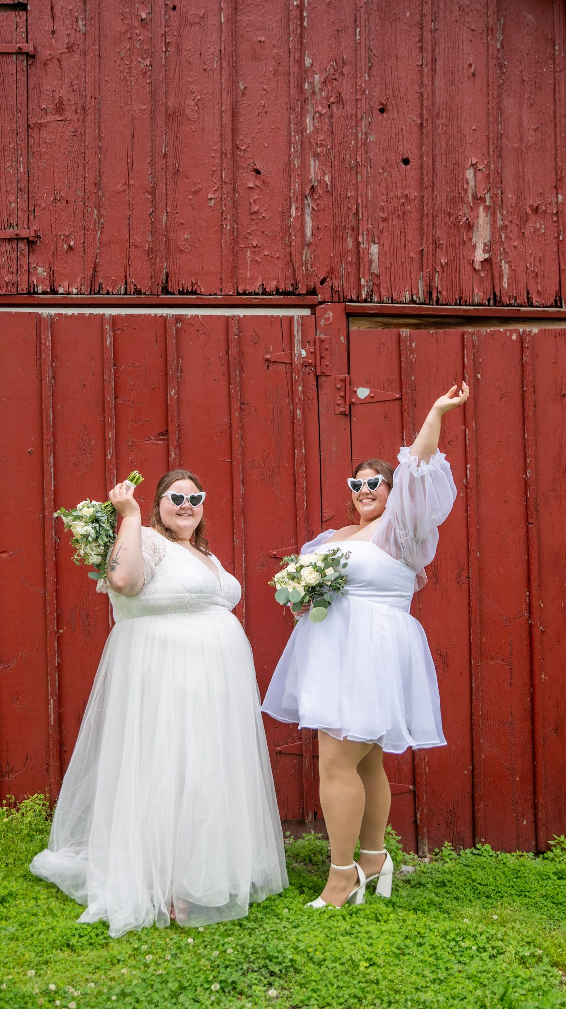 Wedding party in white dresses poses playfully against rustic red barn wall.