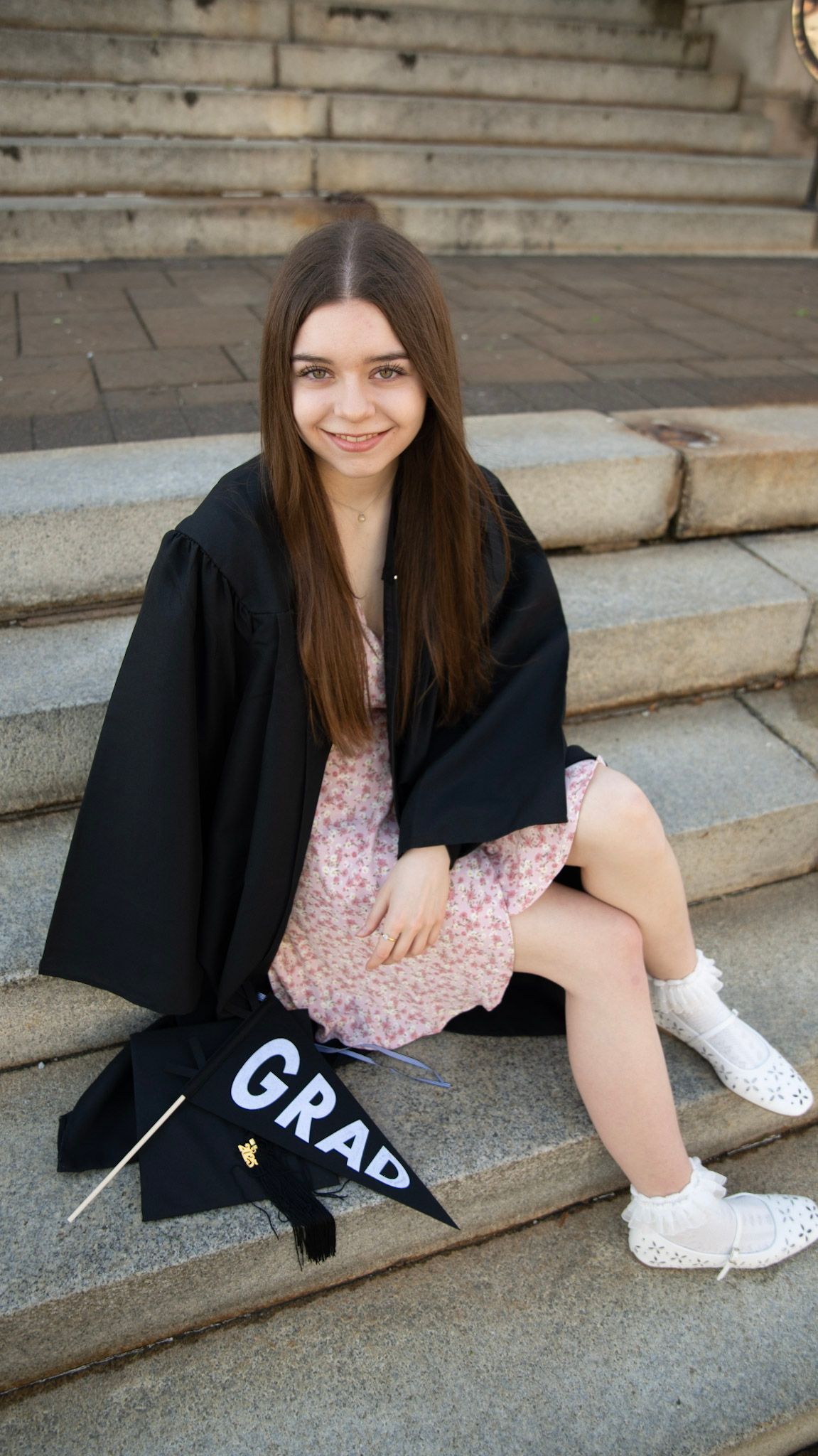 Graduate in a pink floral dress and black gown sits casually on concrete steps.