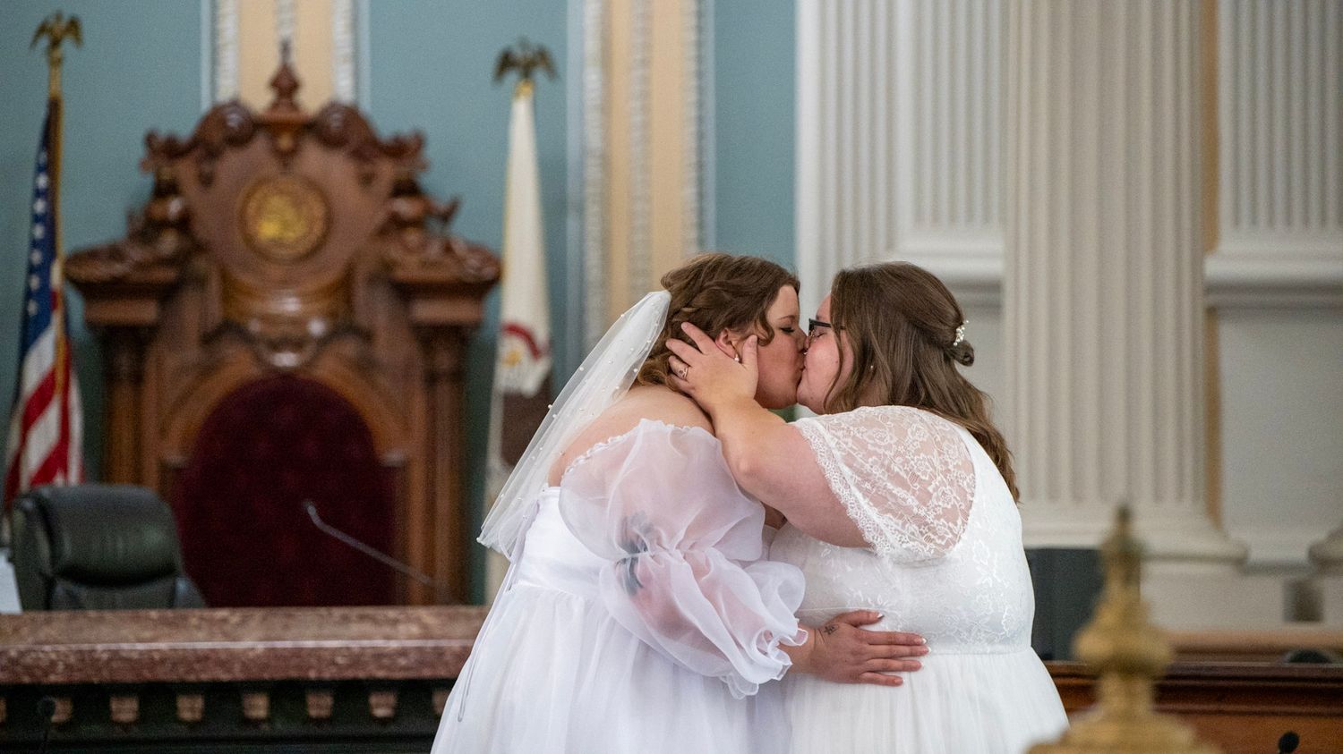 Couple in white dresses embracing during courthouse wedding ceremony.
