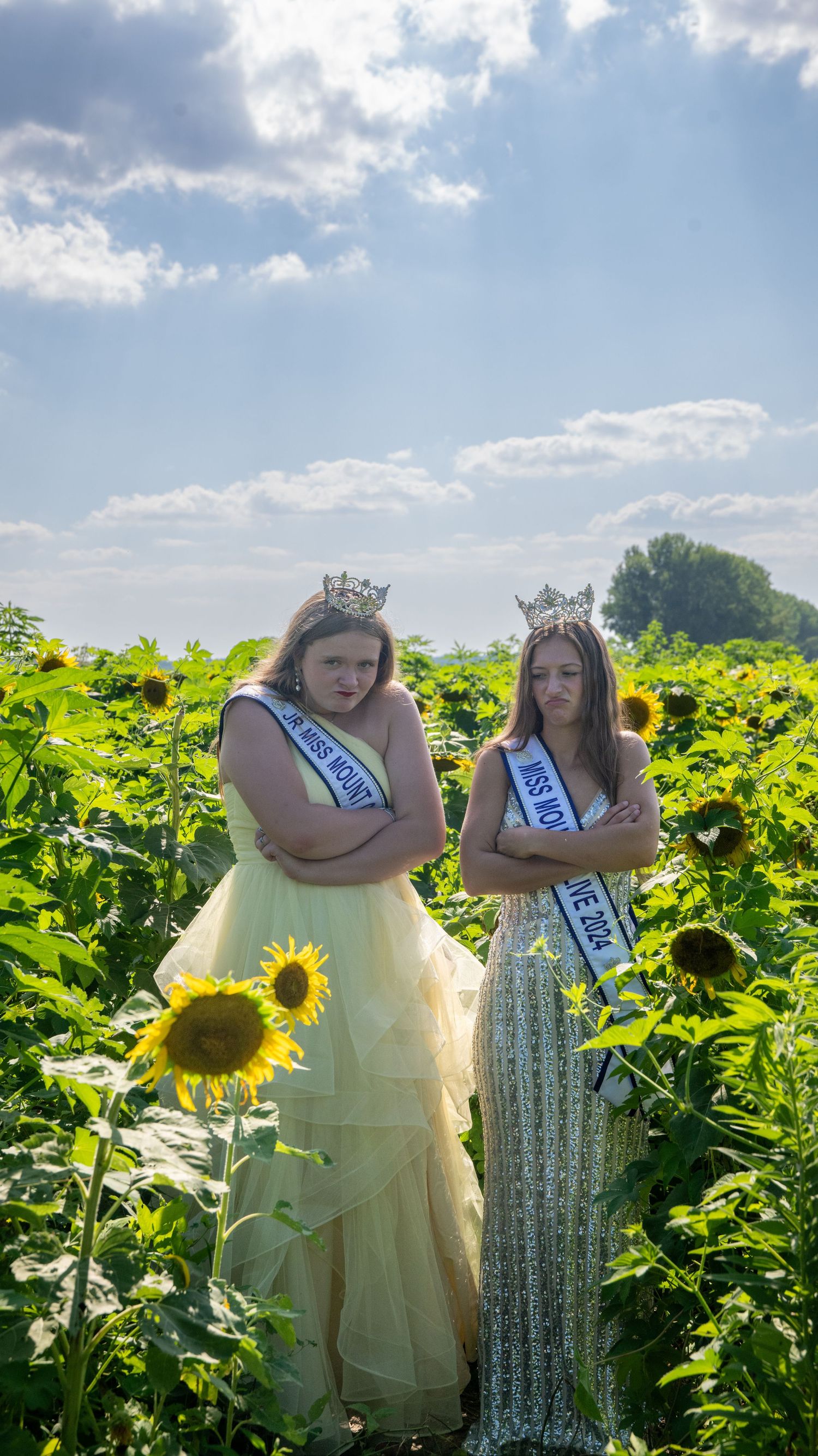 Two pageant winners pose in a sunflower field wearing sashes and crowns.