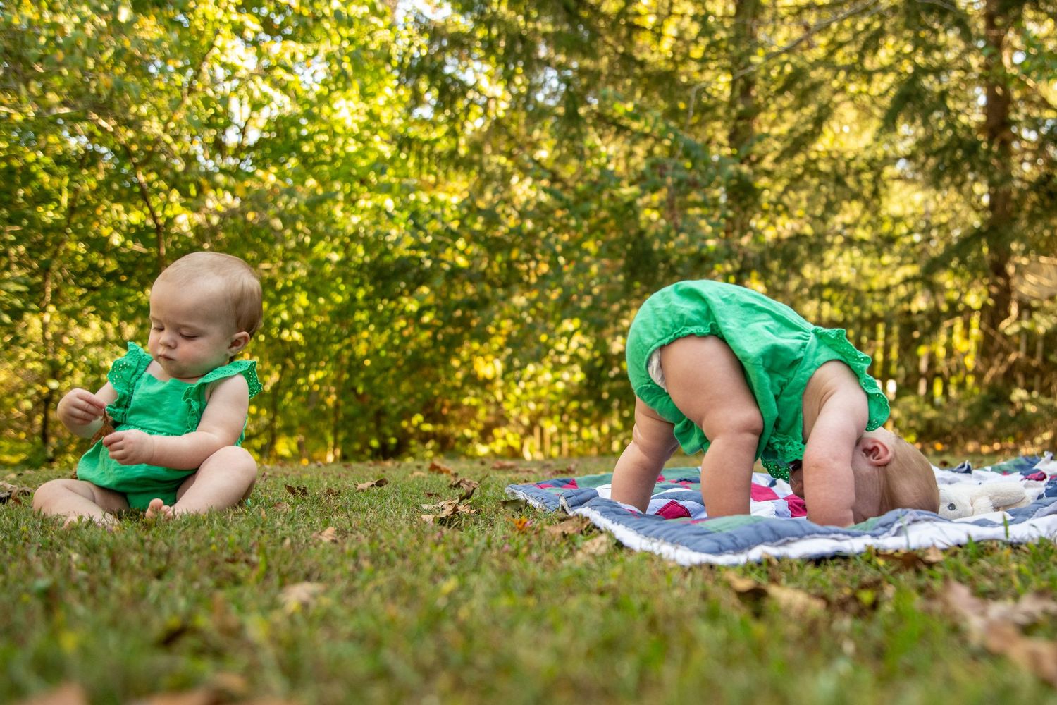 Two babies in matching green outfits playing on blanket in sunlit garden setting.