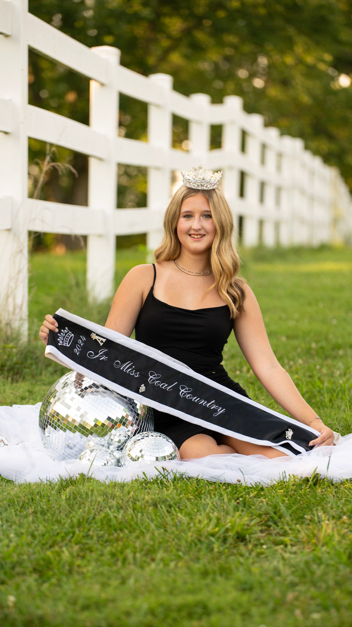 A photo session in a grassy field with a white picket fence featuring a black dress and sparkly accessories.