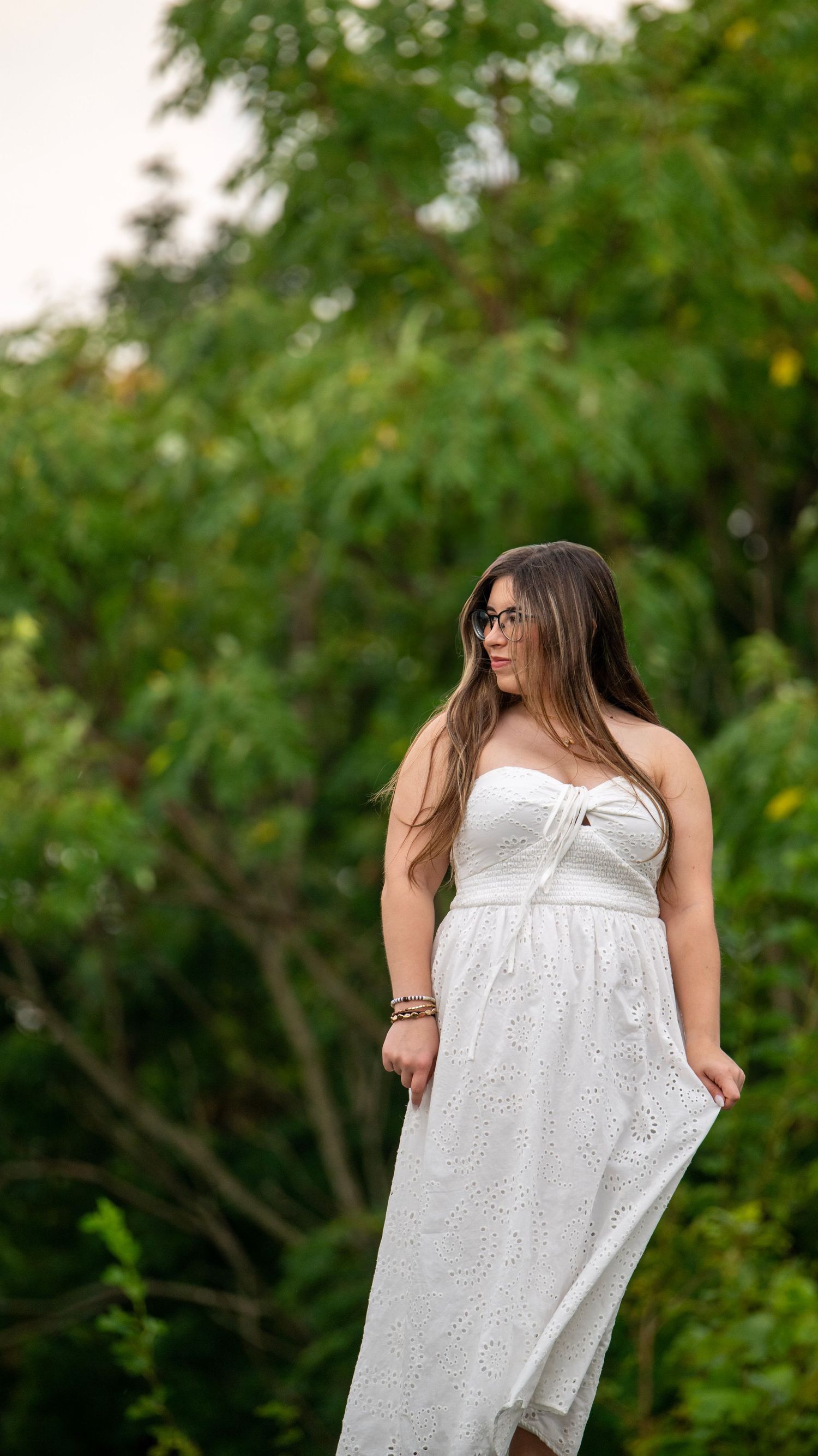 A person wearing a white eyelet dress poses against a verdant natural backdrop.