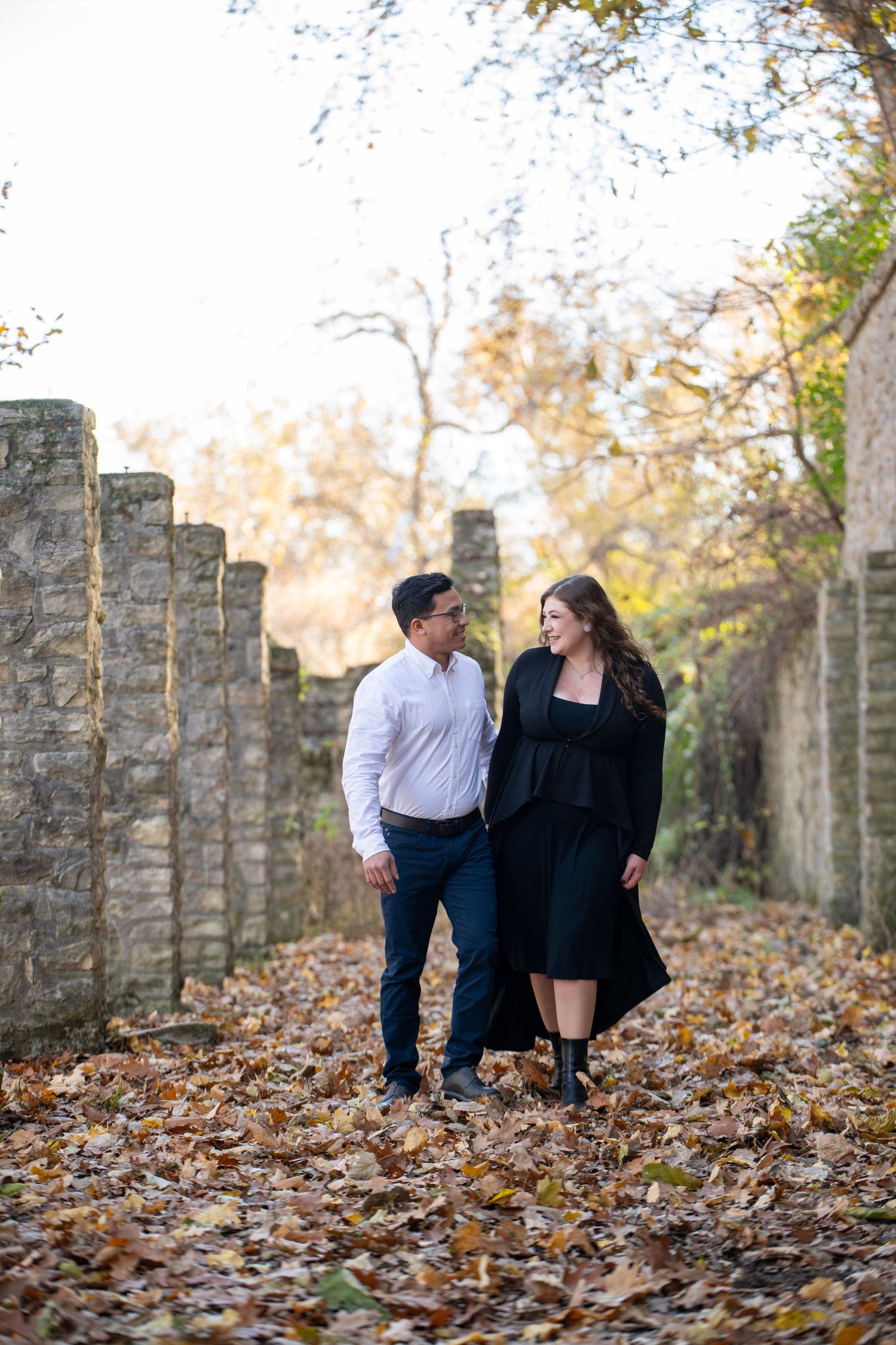 Couple strolls together among autumn leaves beside historic stone colonnade.