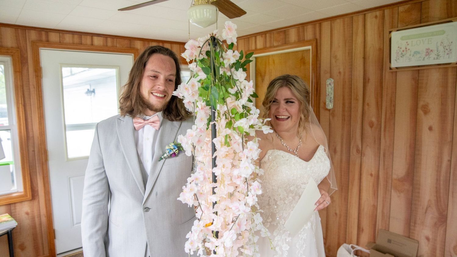 A couple stands together in a rustic wood-paneled room with pink floral decorations for their wedding day.