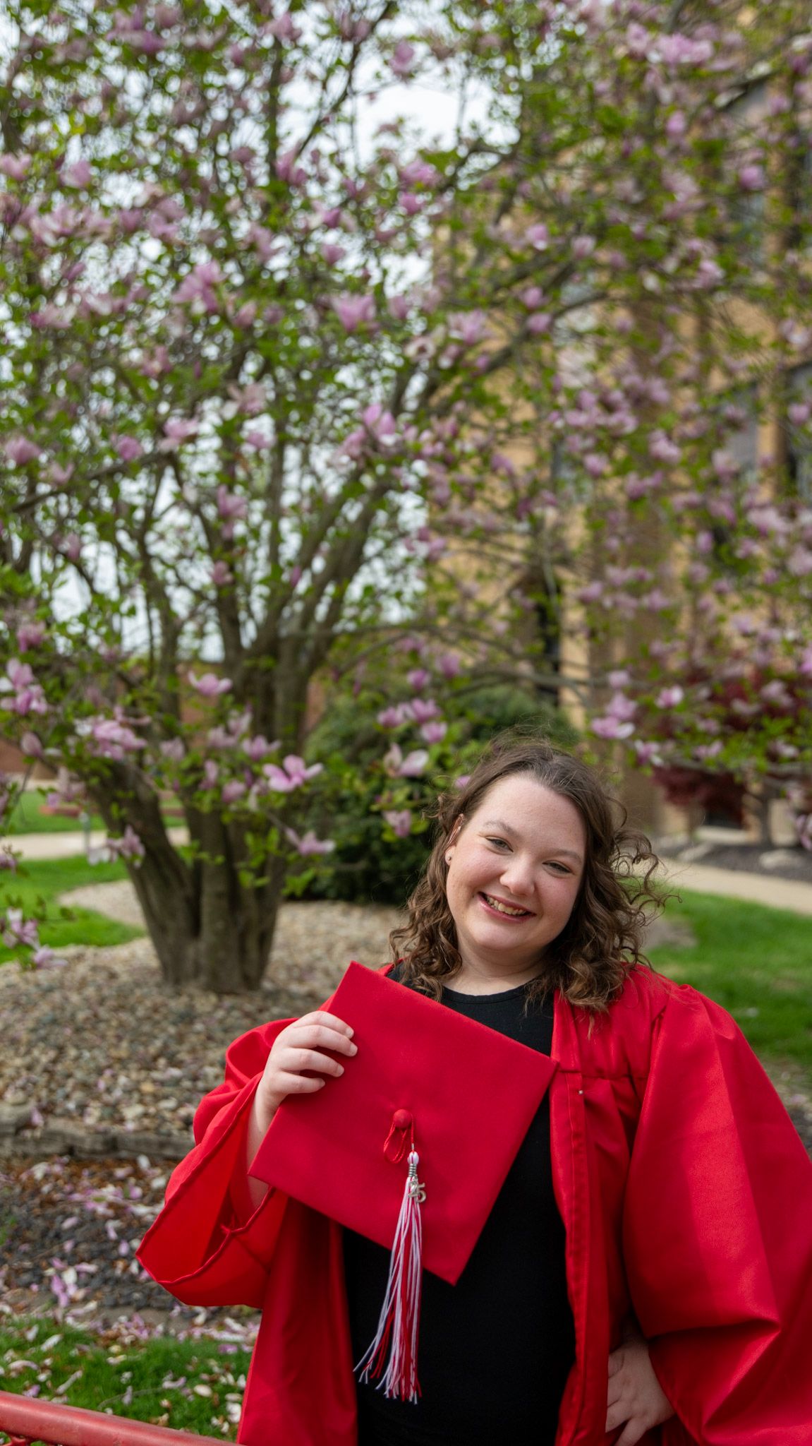 Graduate in red gown poses with diploma against flowering trees in spring.