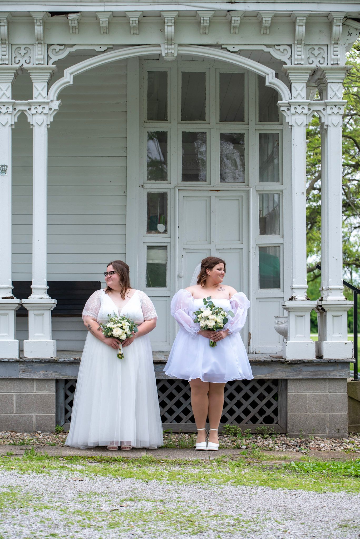 Wedding couple in white dresses posing on vintage porch with ornate white trim.