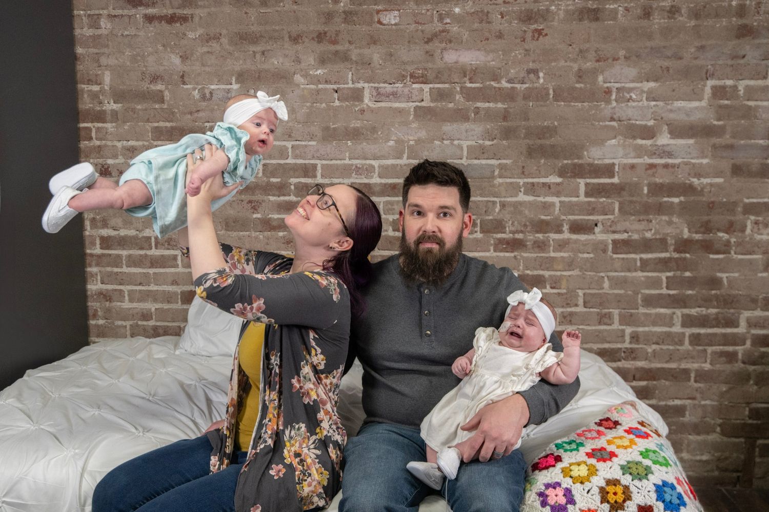 A family poses on a brown leather couch against a rustic brick wall background.