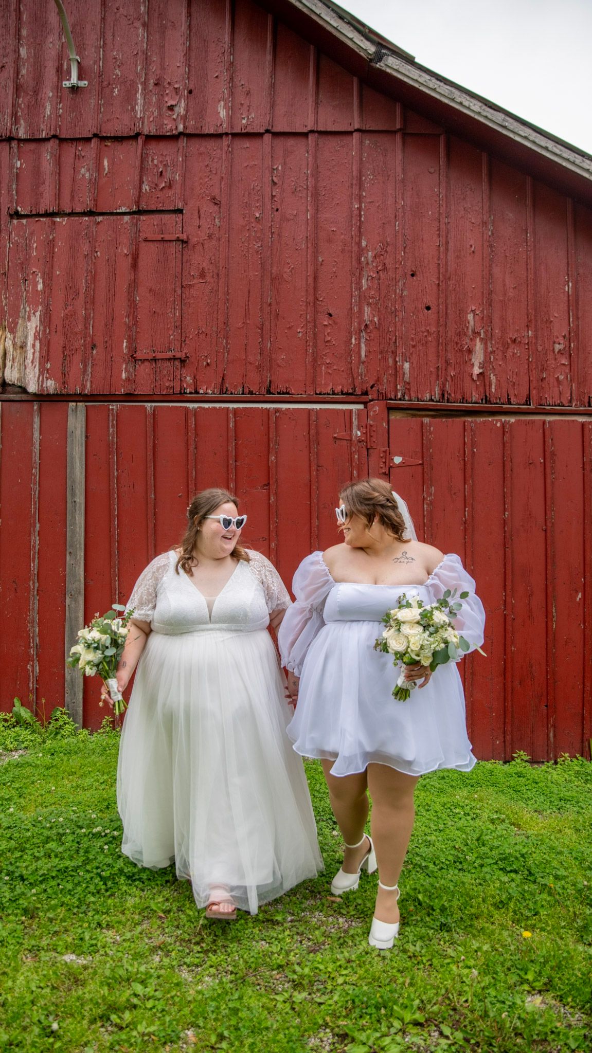 Wedding party poses in white dresses against a rustic red barn backdrop.