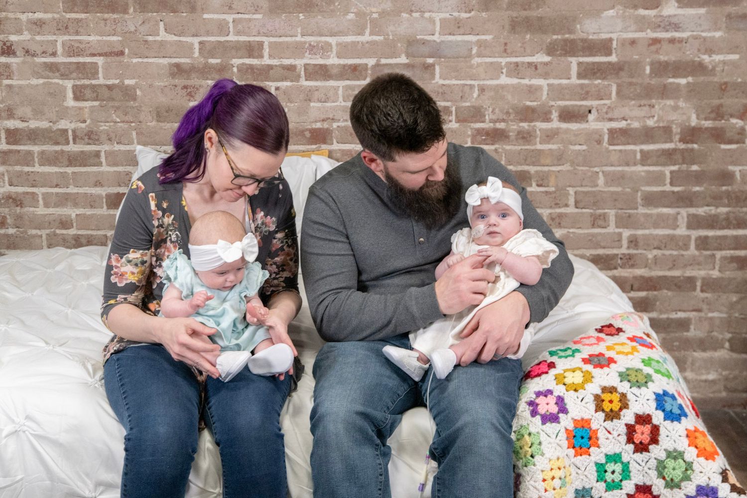 Family portrait with twins on colorful quilt against exposed brick wall background.