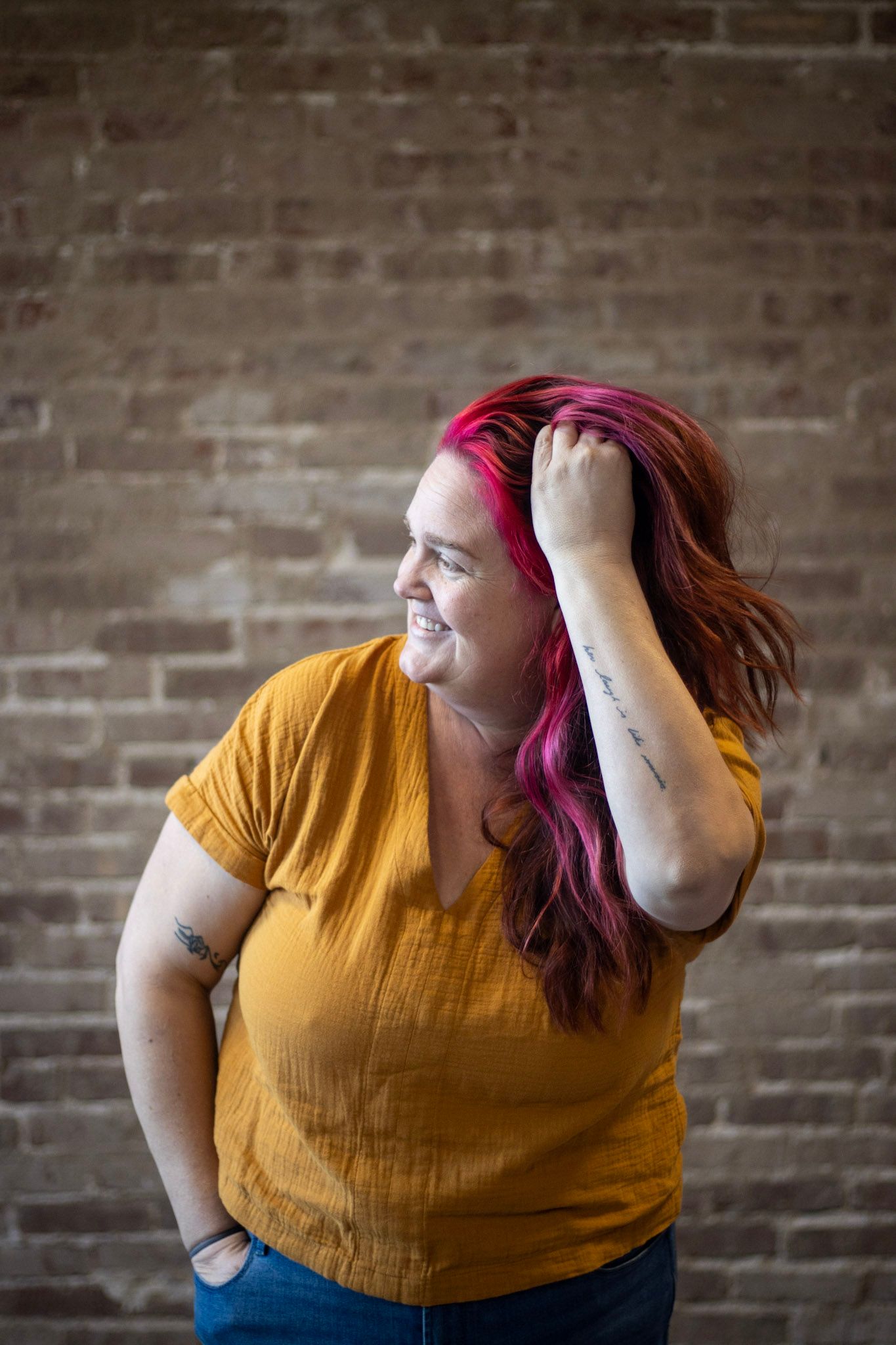 A person in a mustard yellow top poses expressively against an exposed brick wall background.