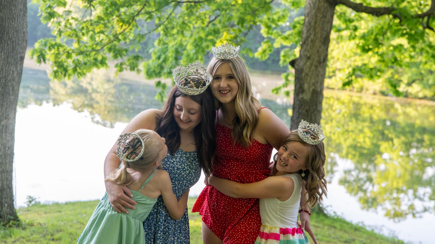Pageant contestants gather for group photos by a lake on a sunny day.