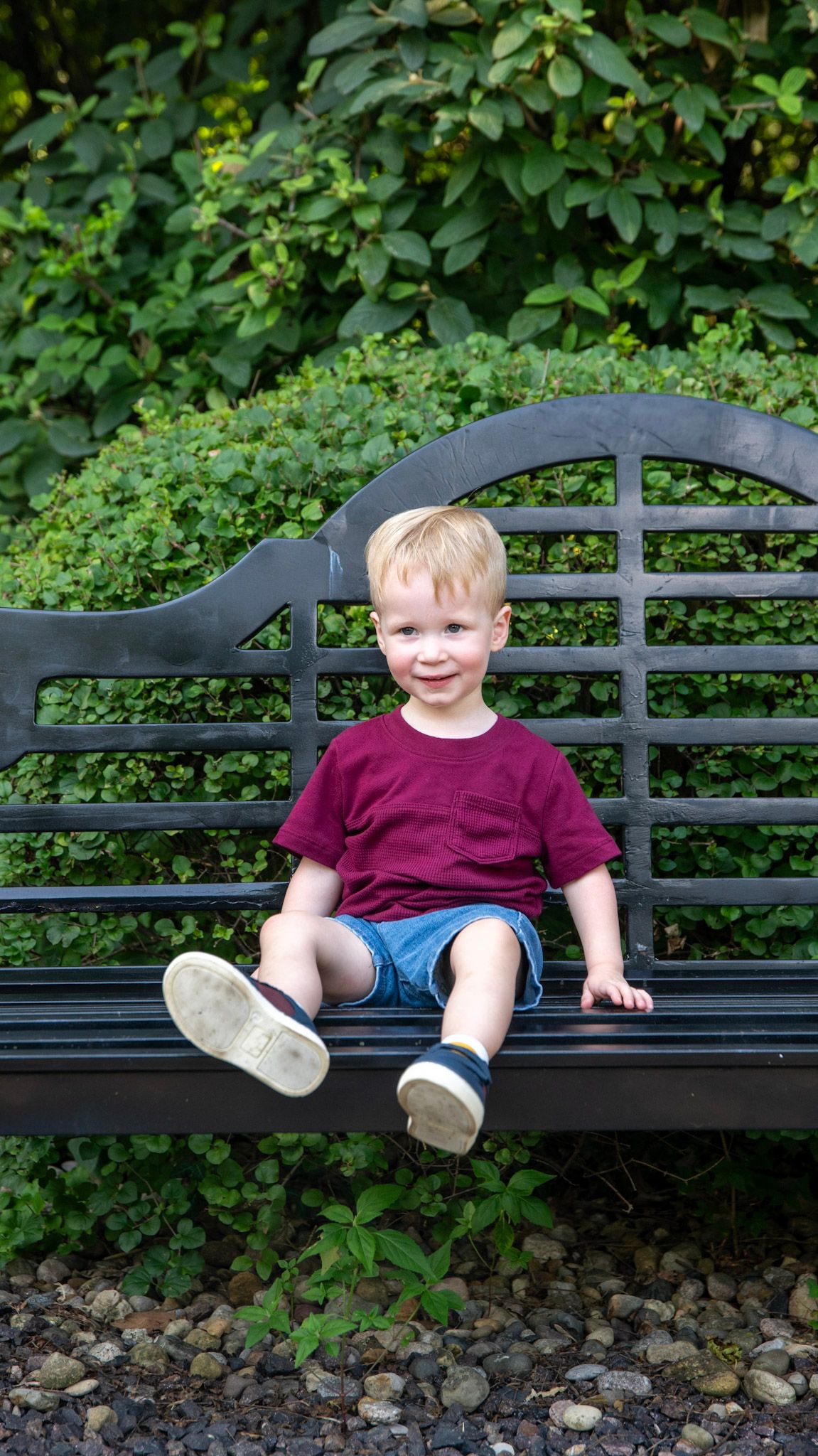 Children sit on a black metal bench surrounded by lush green foliage.