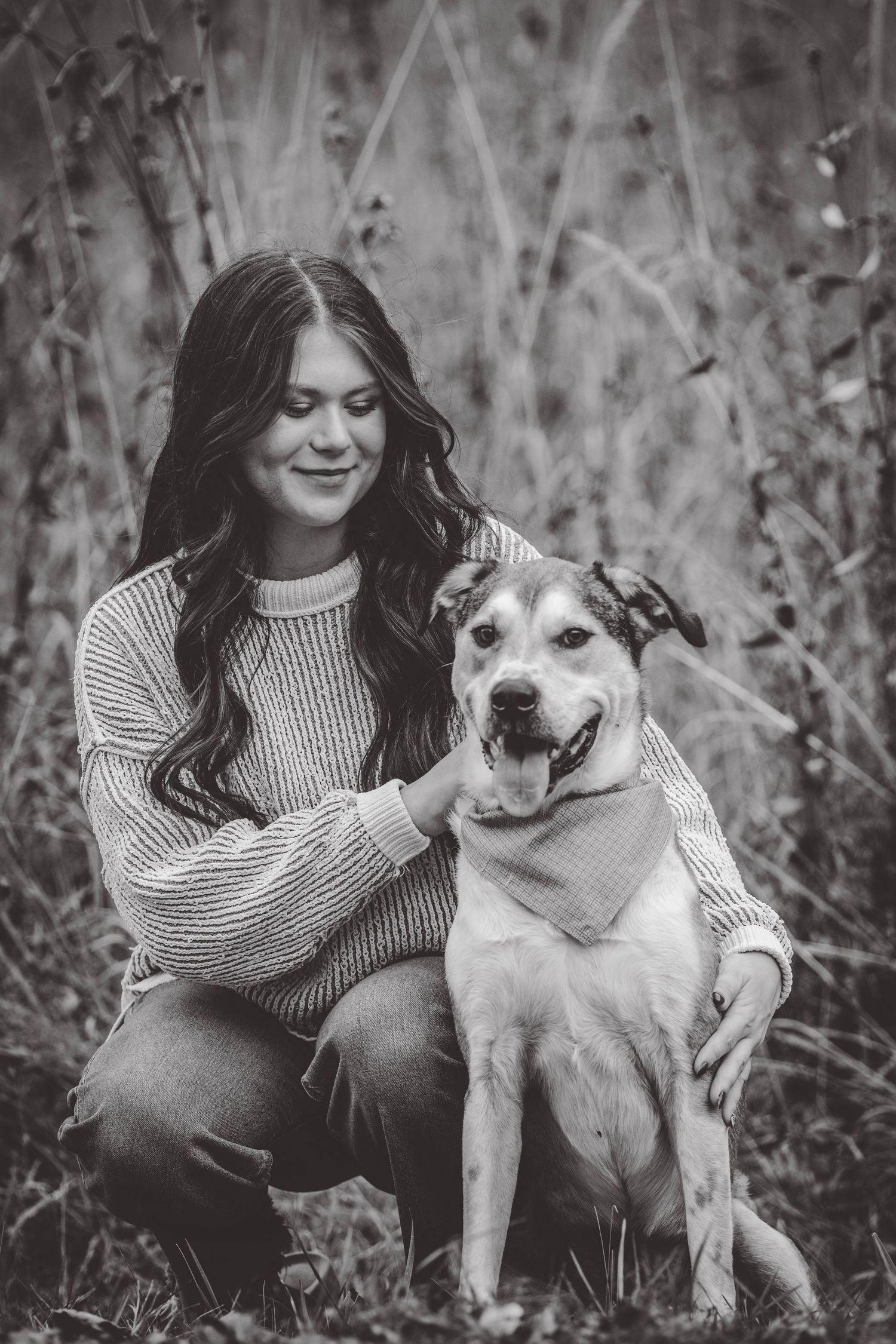 A person in a striped sweater sits with their mixed breed dog in a black and white outdoor portrait.