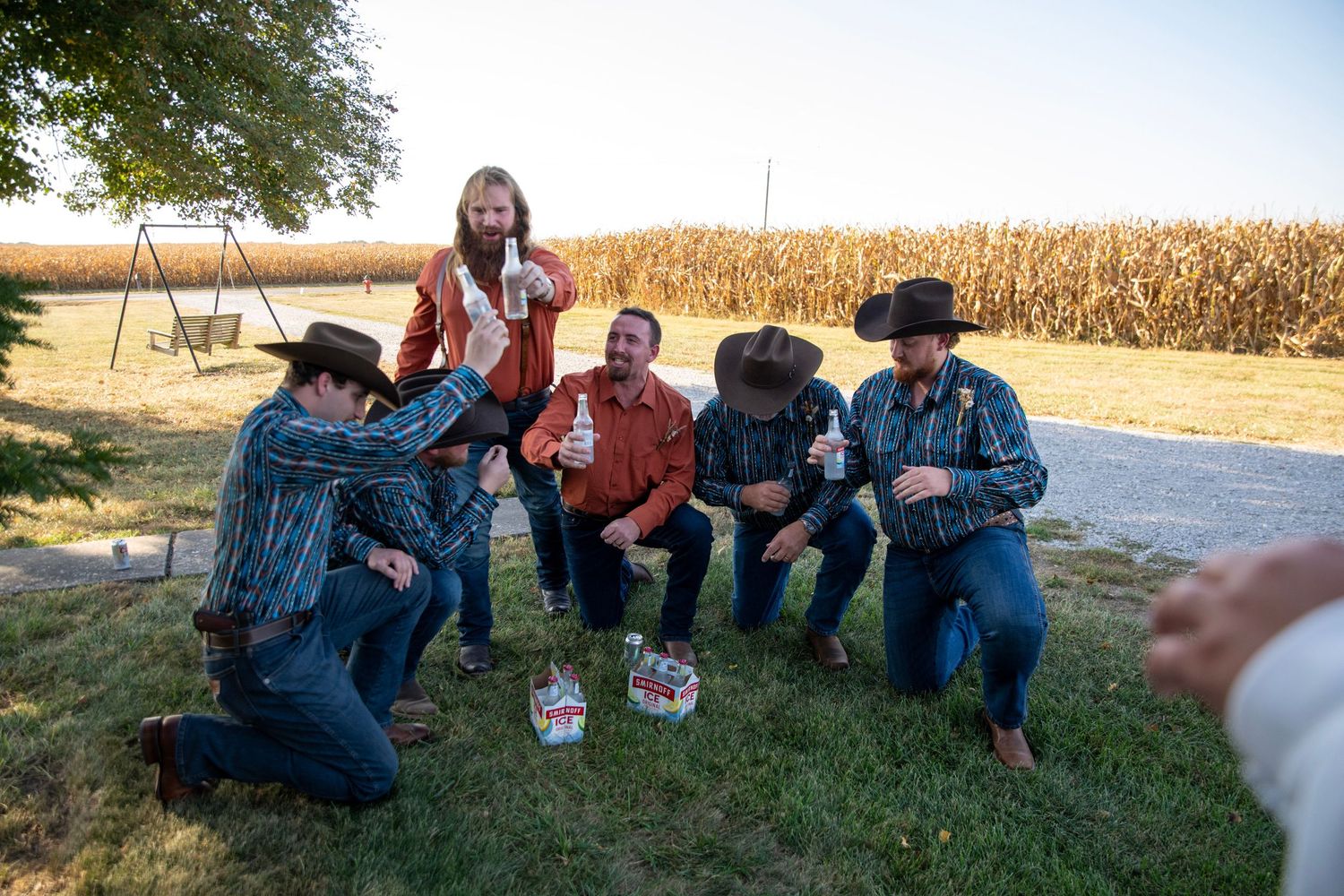 Friends in casual western attire socialize together on grass near a cornfield.