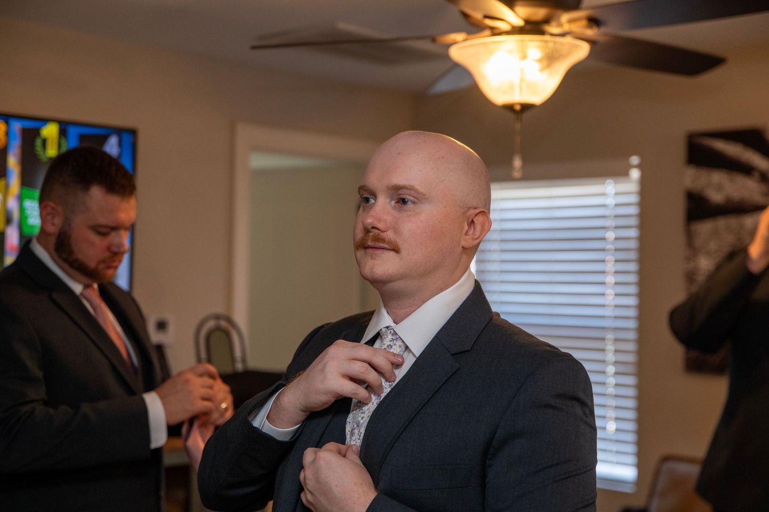 People in formal suits gathered in room with ceiling fan and window light.