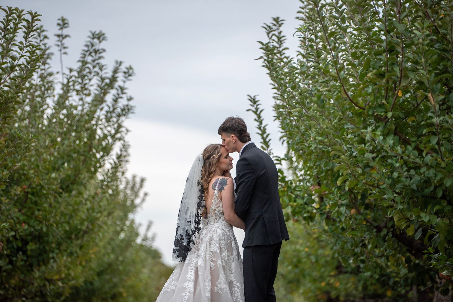A romantic wedding moment captured between trees in a lush orchard setting.