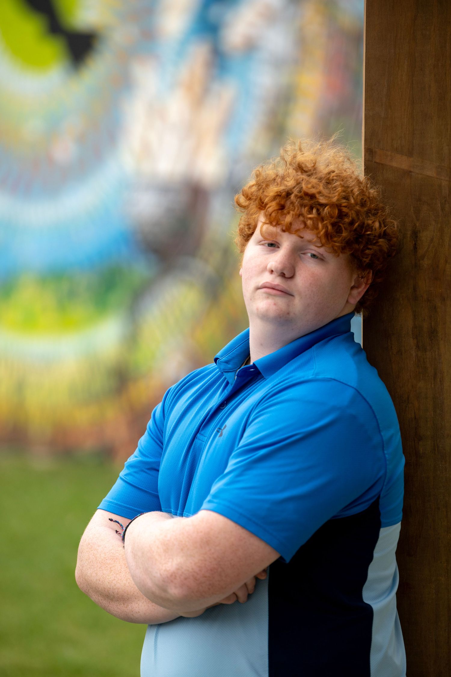 Young person in bright blue shirt posing against wooden pillar in outdoor setting.