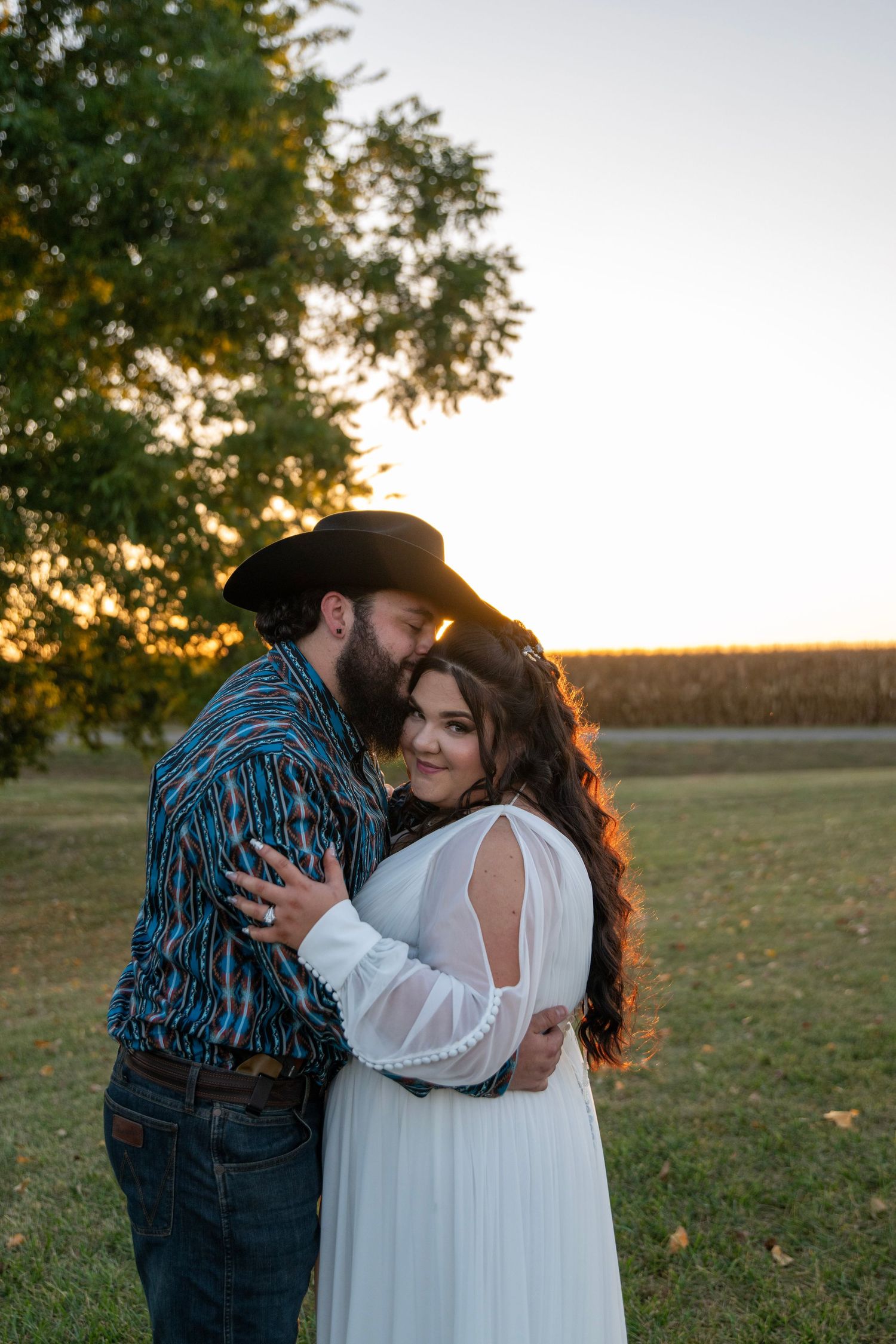 Couple embraces in white dress and western wear during golden hour in rural field.