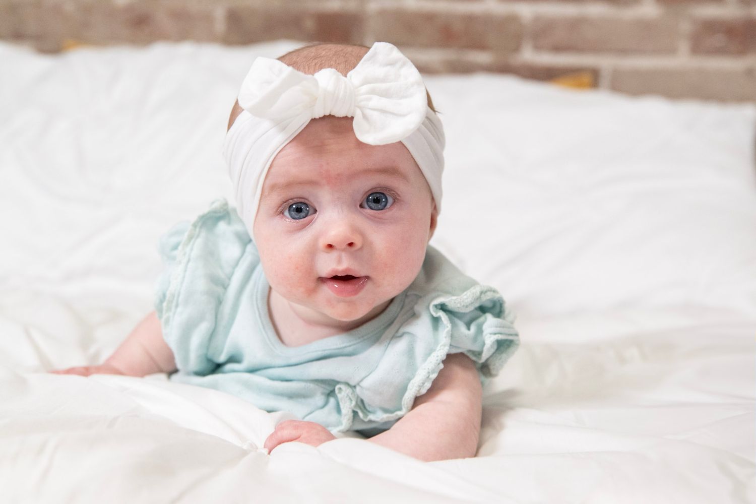 Adorable baby wearing mint green dress and white bow headband smiles on white bedding.