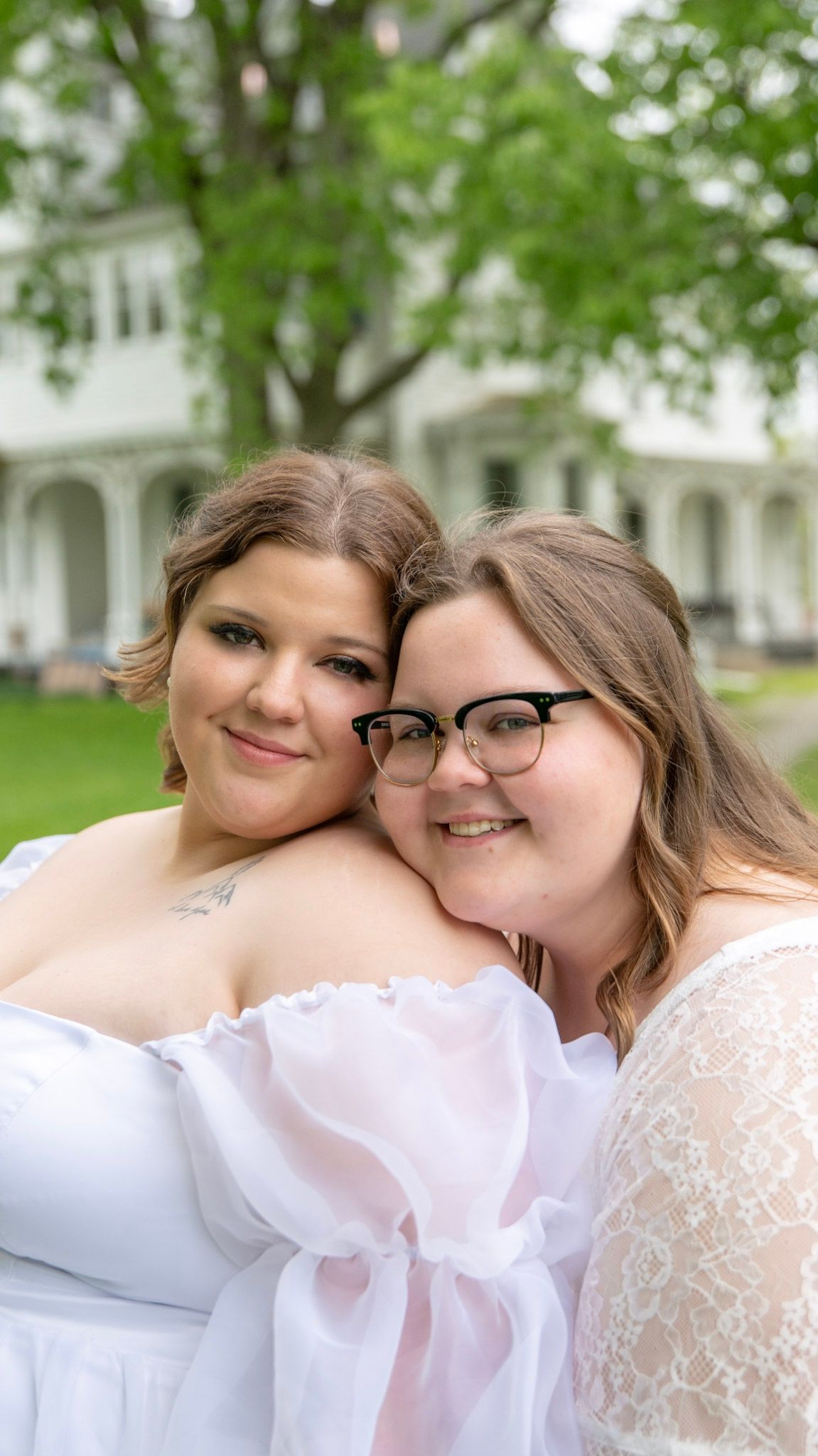 A couple in white dresses shares an intimate moment outdoors with green trees in background.