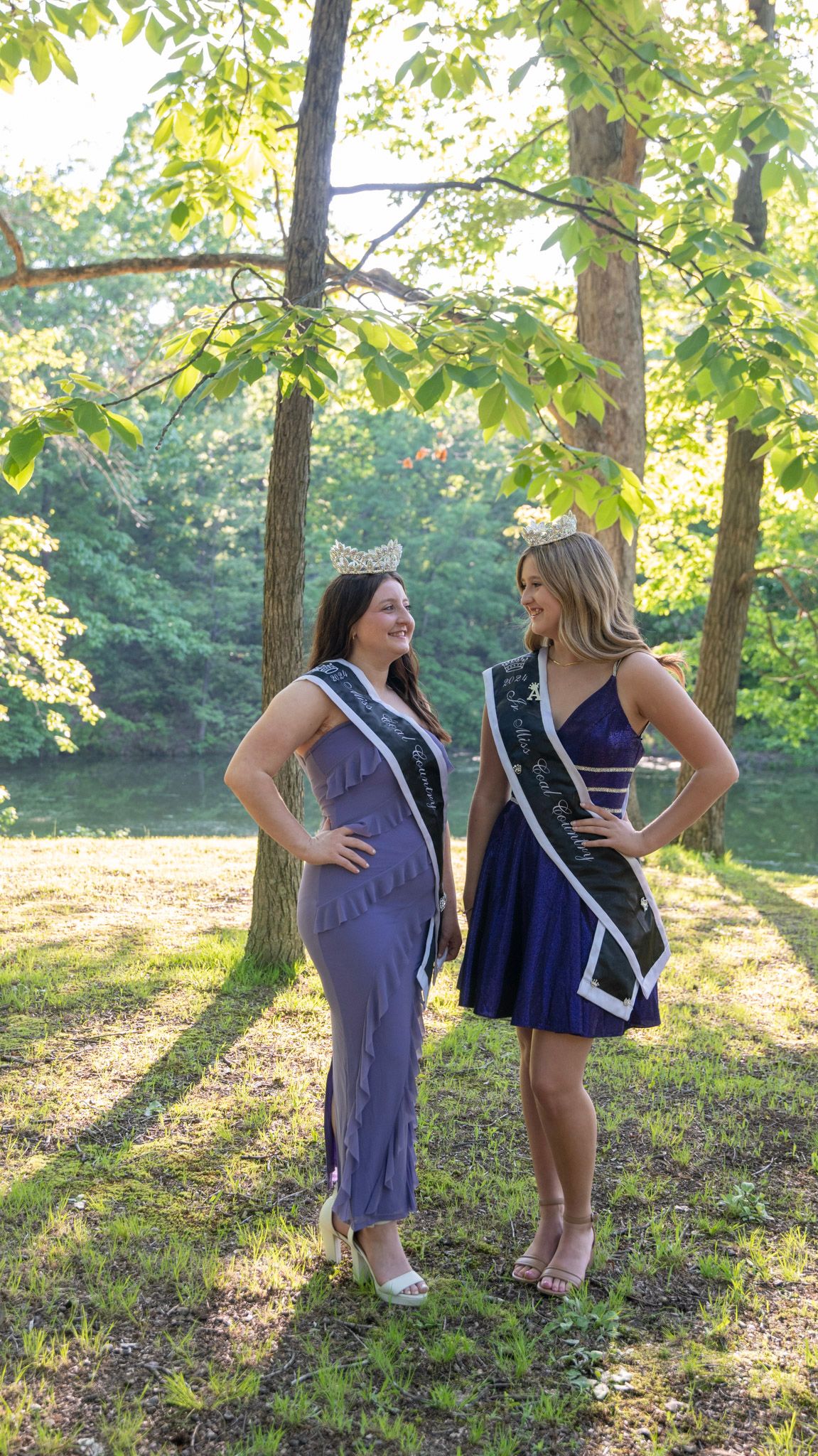 Two pageant contestants share a laugh while posing in formal dresses outdoors.