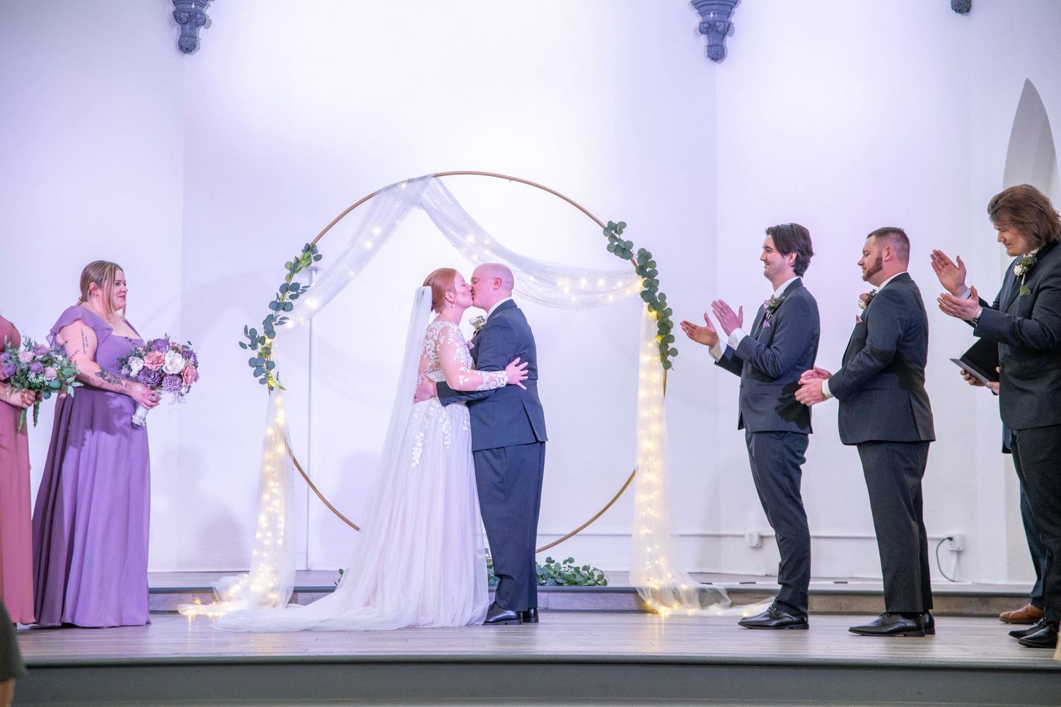 Wedding party stands at altar under circular arch with purple-clad bridesmaids and groomsmen in dark suits.