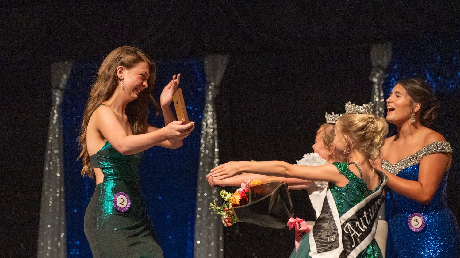 Pageant participants in evening gowns interact on stage during a competition.