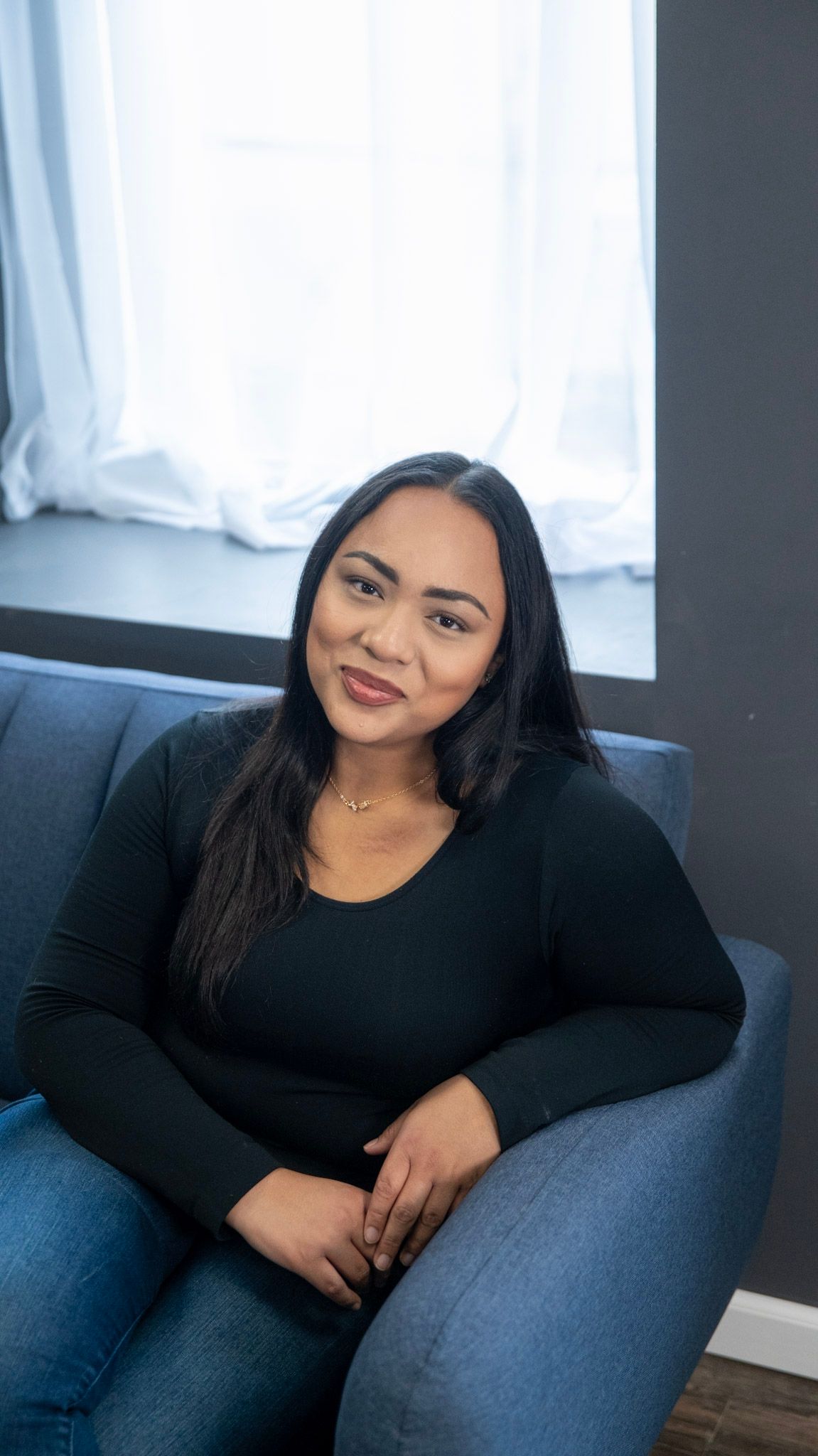 Woman in casual black top posing on grey couch with bright window background.
