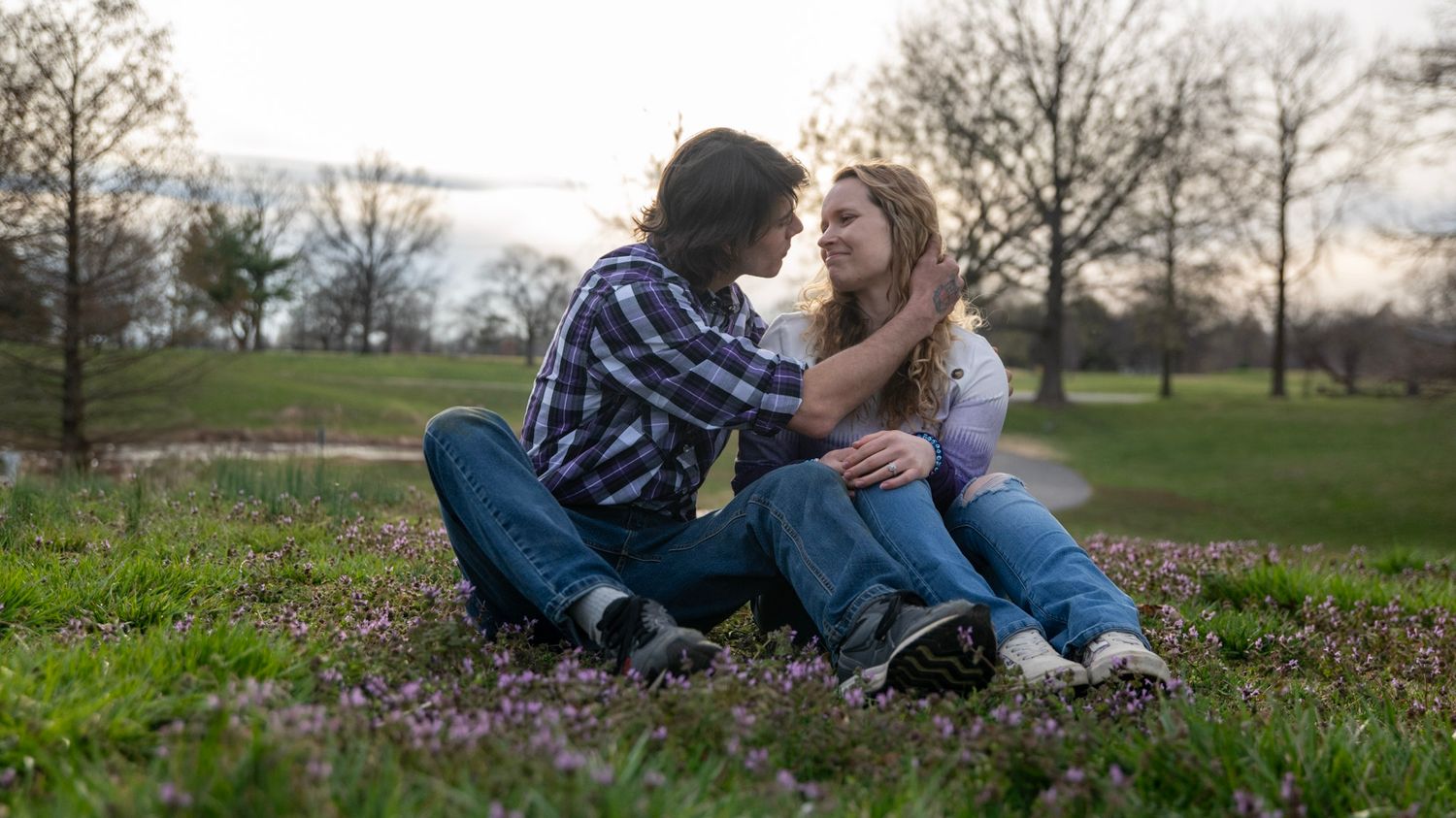 A romantic couple embracing while sitting on grass in a park at sunset.