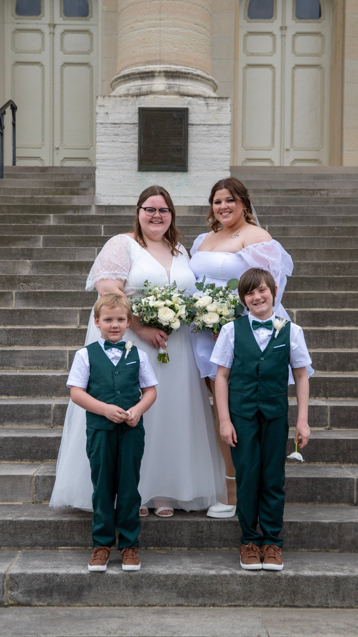 A wedding party poses on church steps with two brides in white dresses and two children in green vests.
