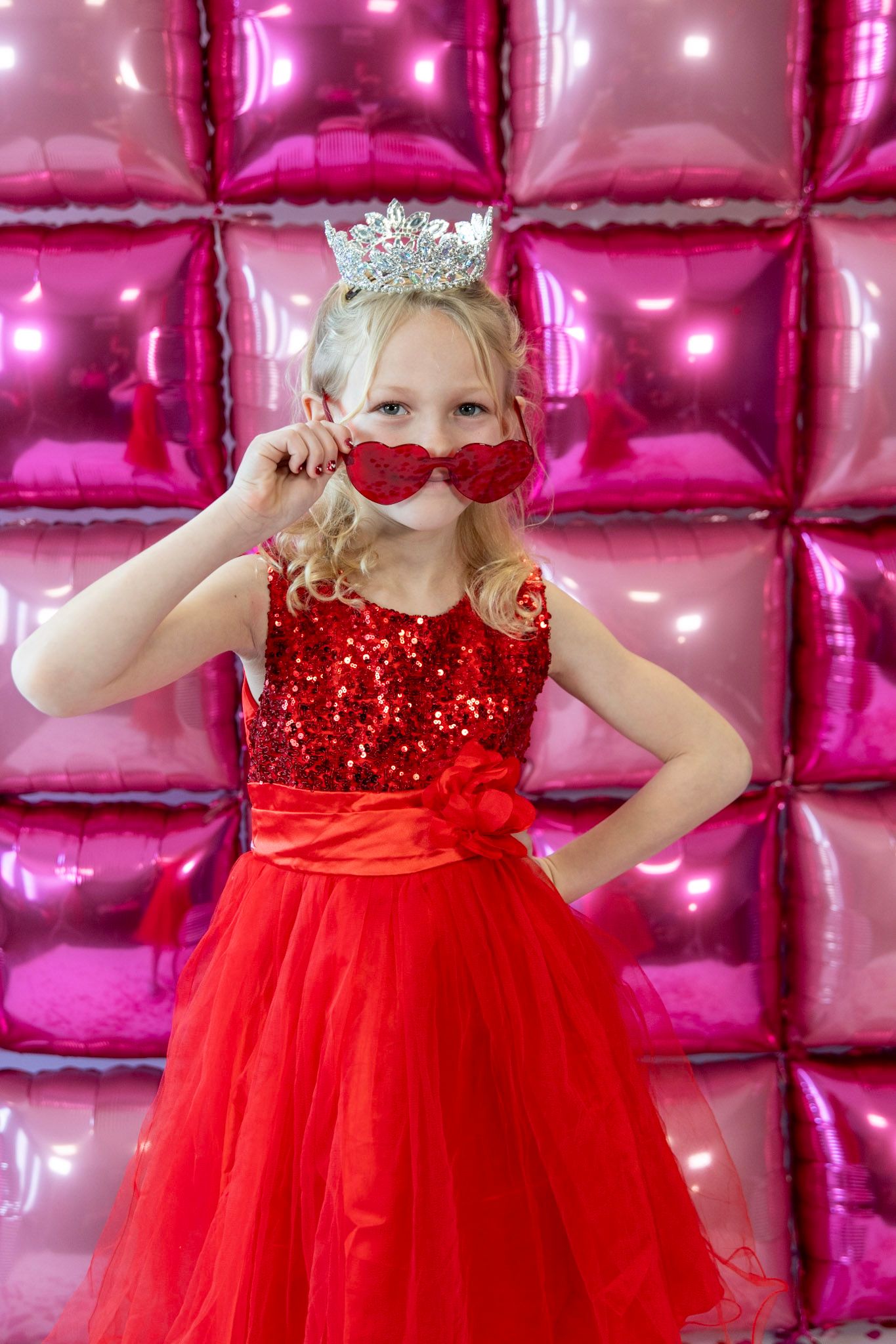 Red tulle dress and crown against shimmering pink balloon wall background.