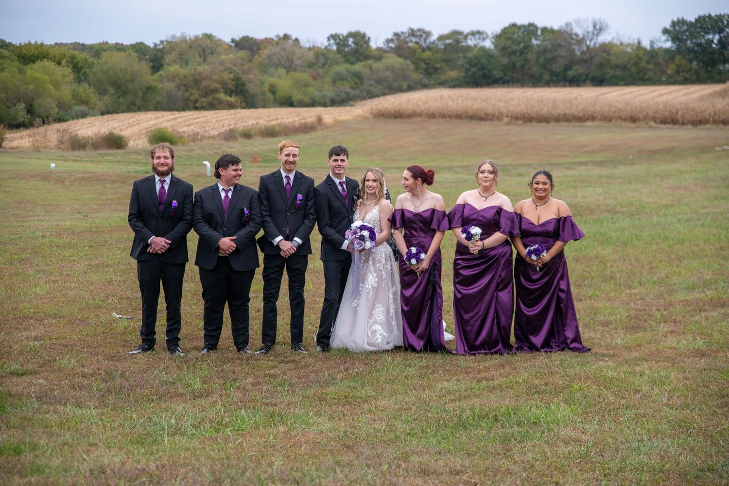 Wedding party poses formally in purple dresses and black suits against rolling countryside landscape.