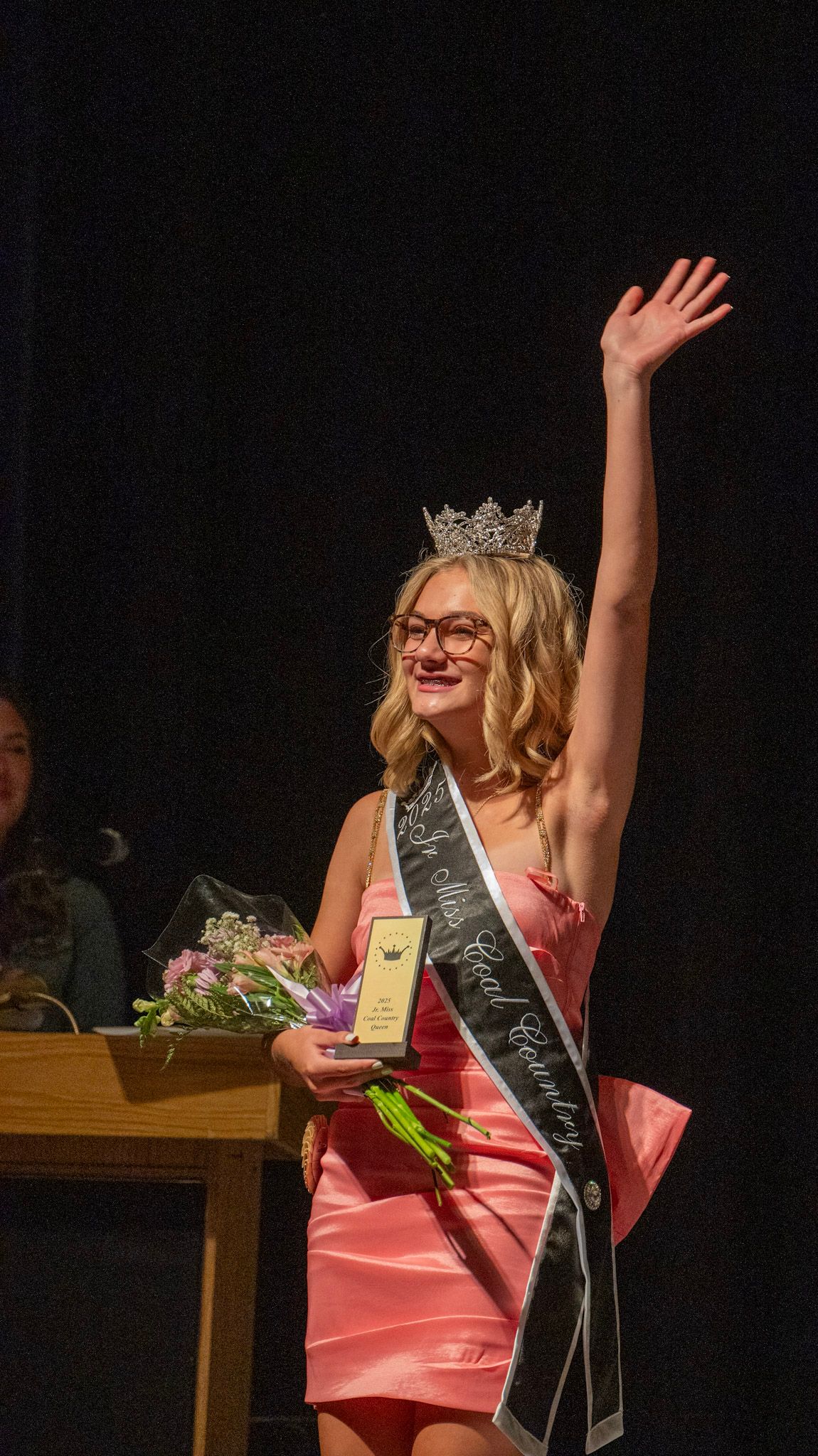 Pageant winner in coral dress and crown waves while wearing sash and holding flowers.