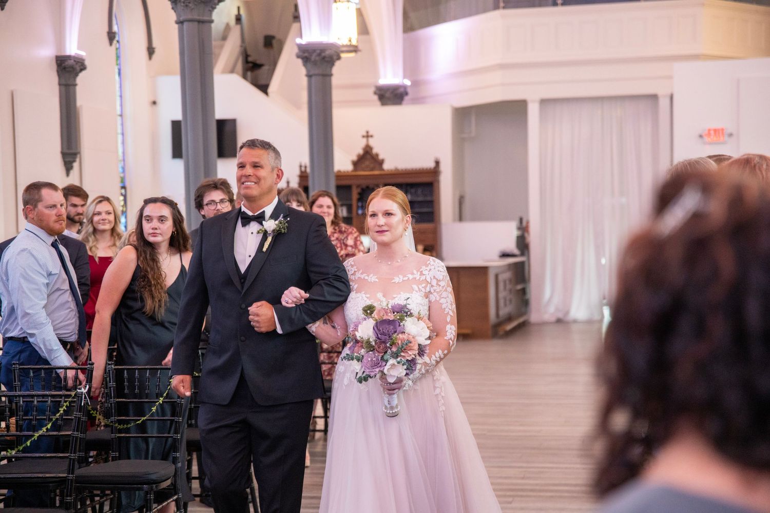 Wedding party and guests gathered in an elegant white church interior.