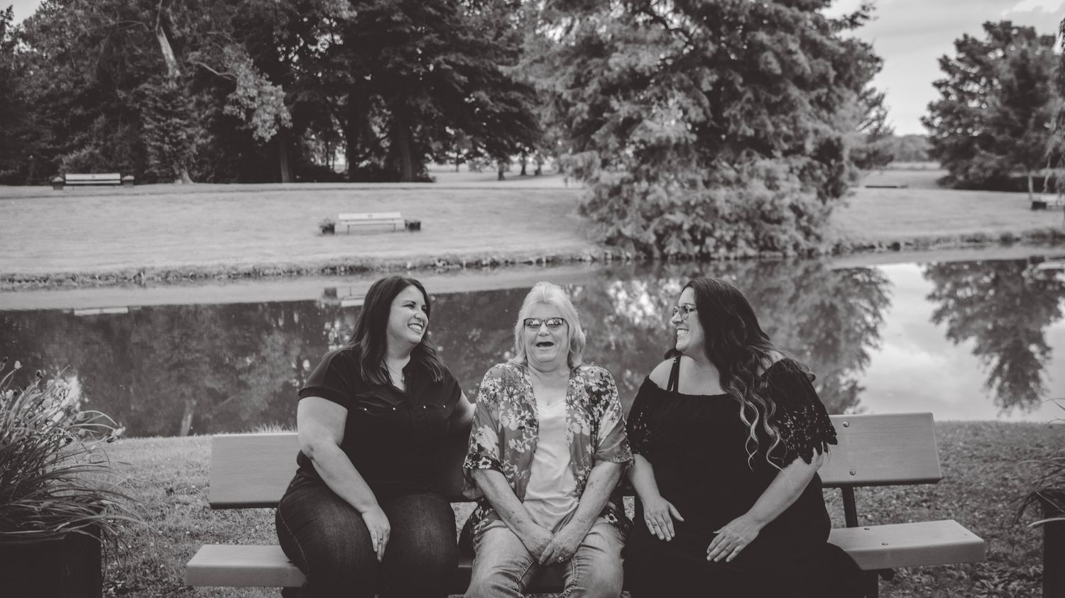 A black and white photo captures three friends sharing laughter on a park bench by water.