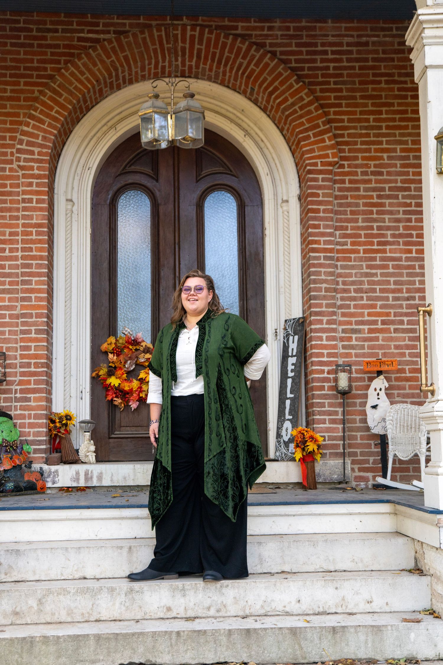 Two people in formal wear stand on the front steps of a classic brick building with fall decorations.