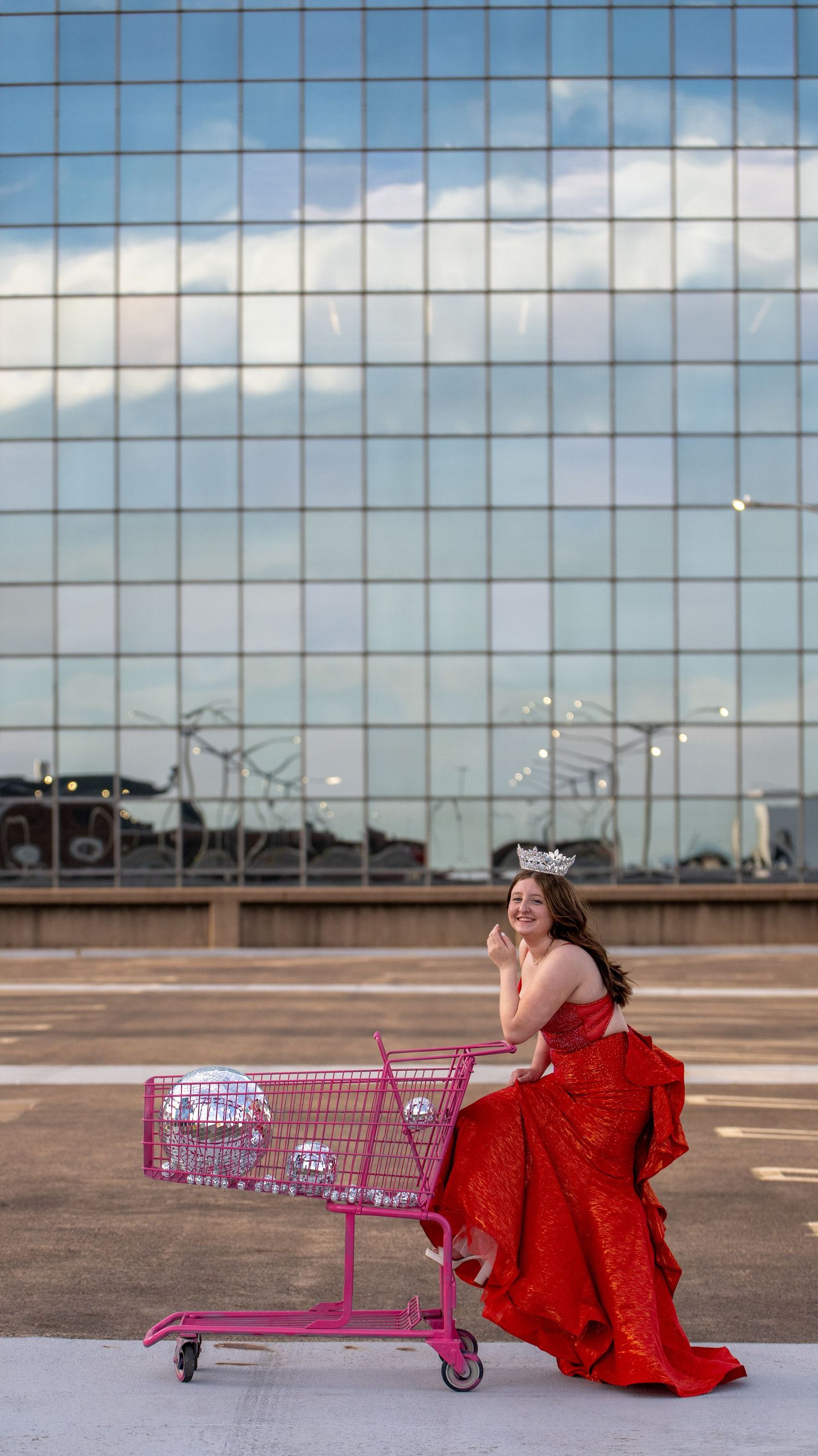 A fashion photoshoot with a red dress and pink shopping cart in an urban parking lot.