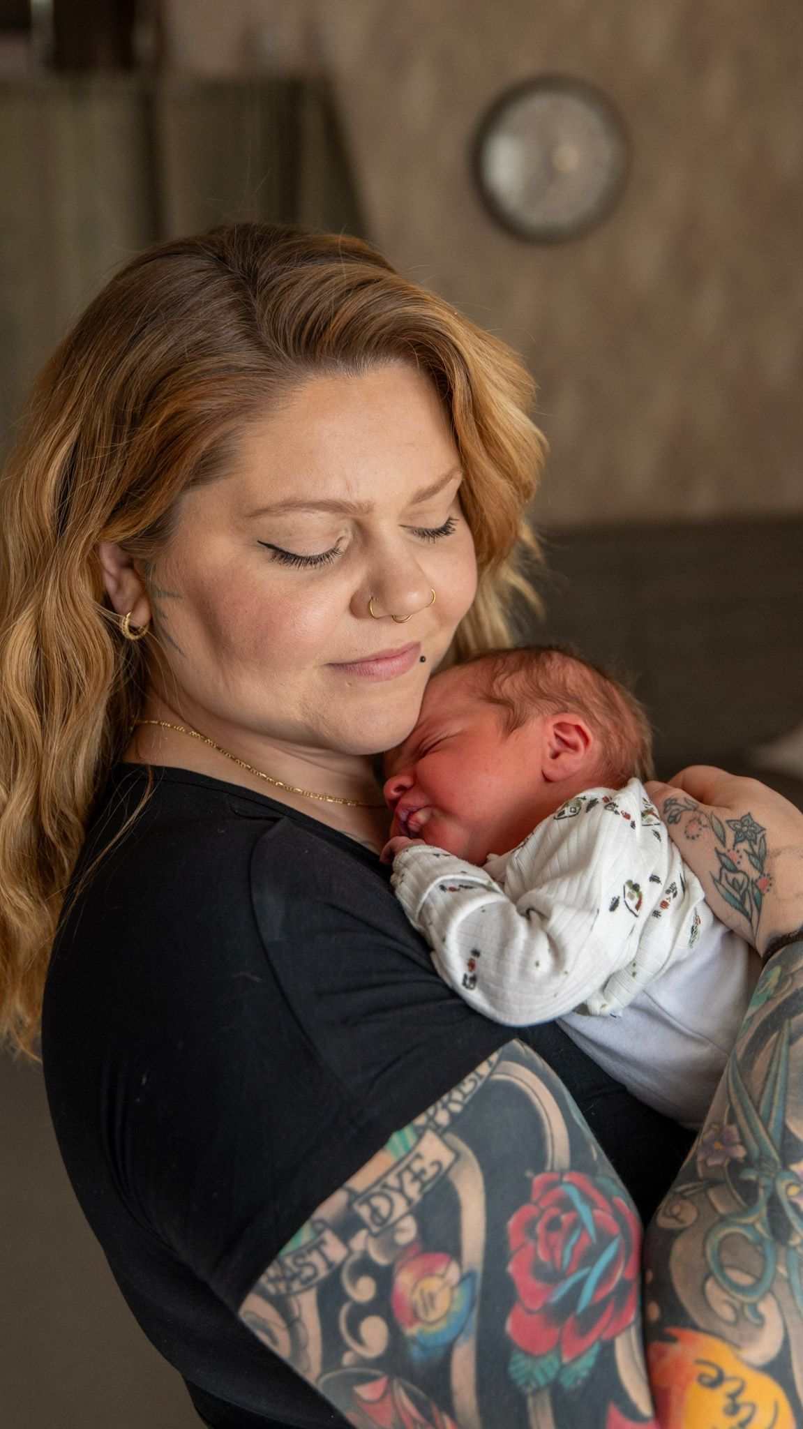 Person with colorful arm tattoos holding and bonding with a newborn baby wrapped in white.