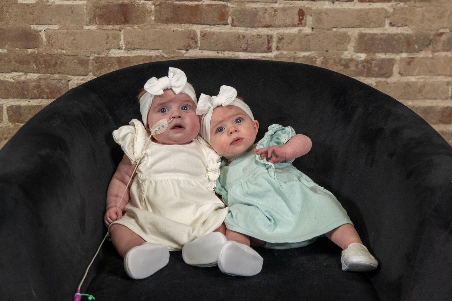 Twin babies wearing matching outfits in cream and mint green sit together on black couch.
