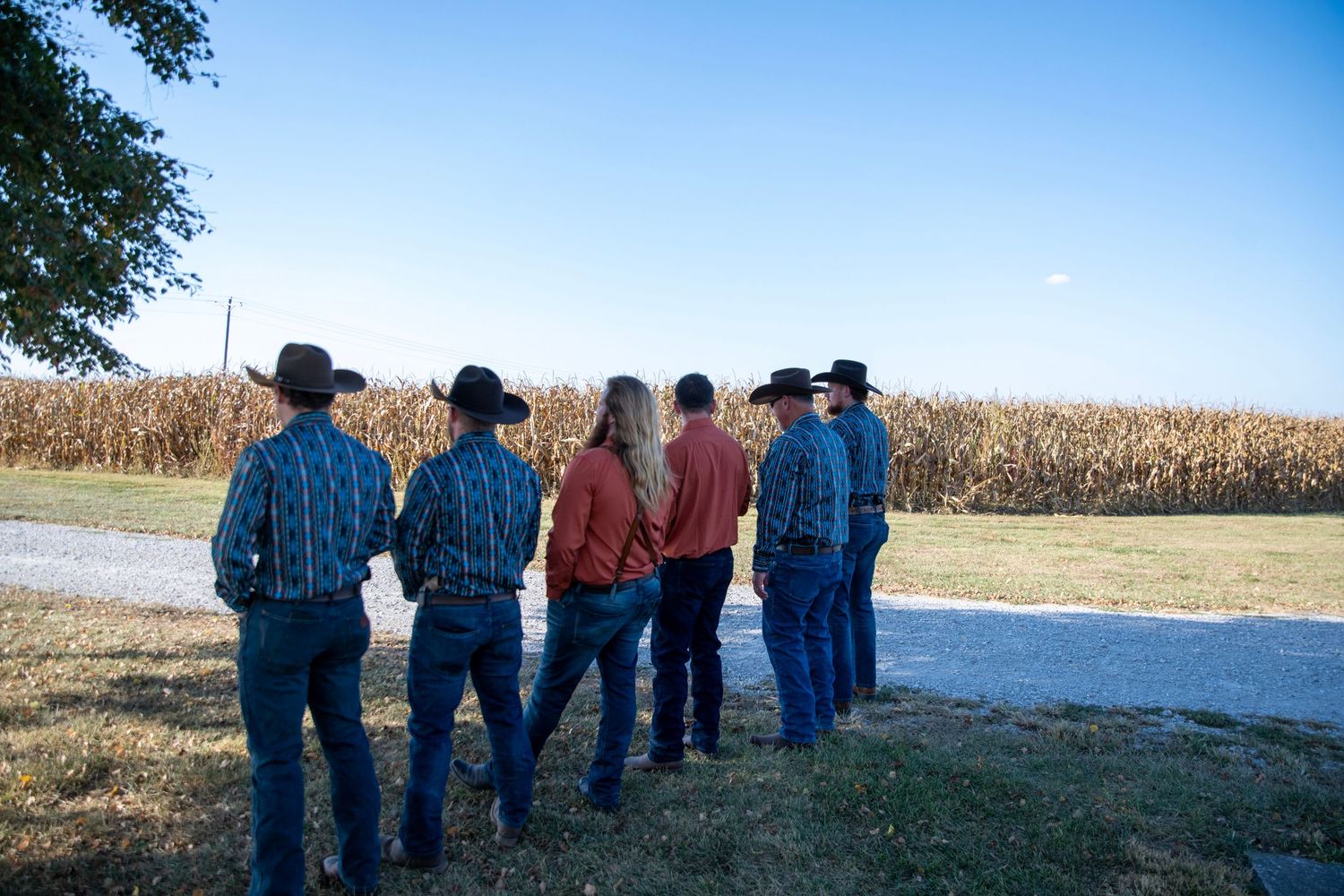 A group of people in cowboy hats and western wear stand together in front of a cornfield at sunset.