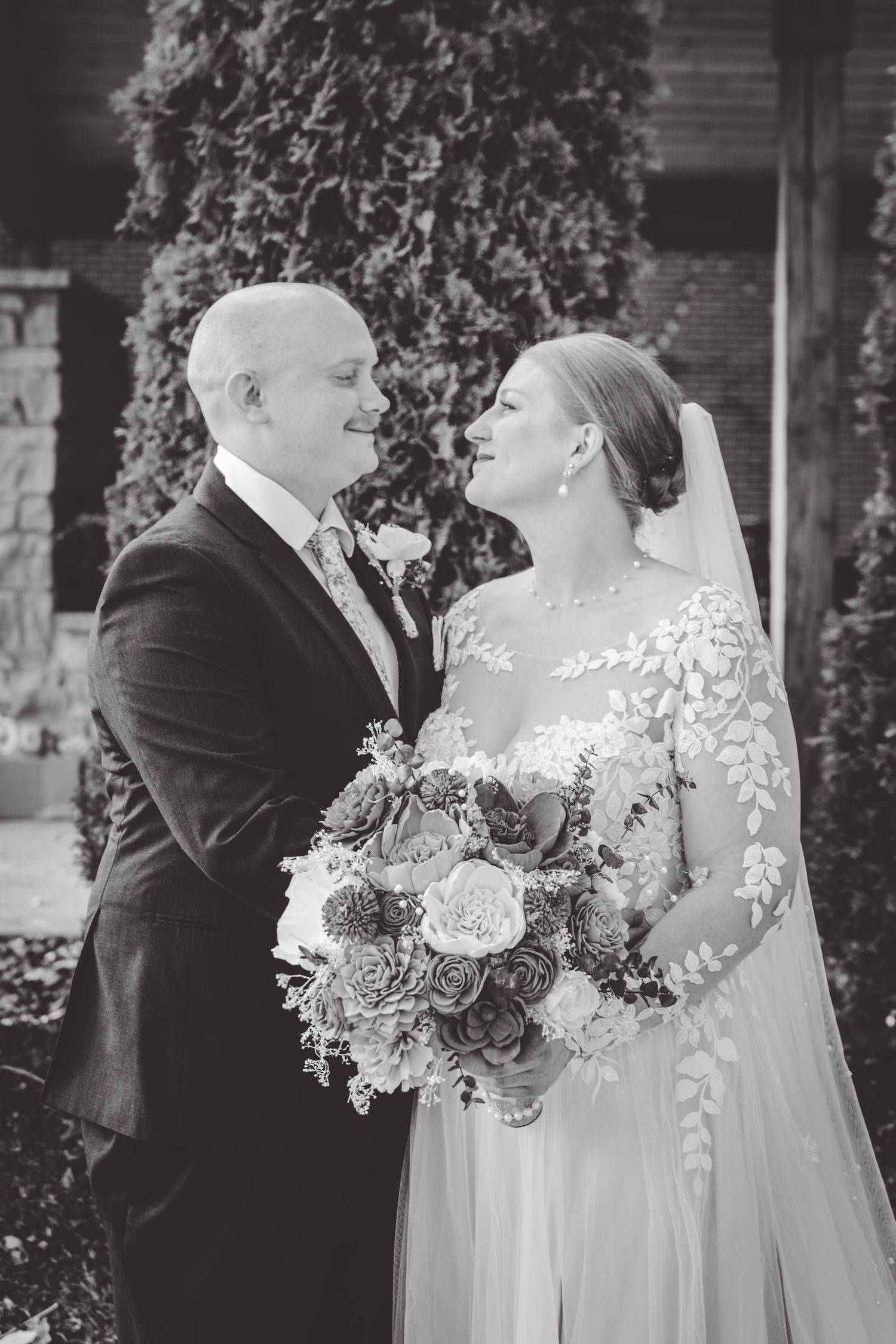 Black and white portrait of wedding couple with vintage-inspired bouquet against brick wall.